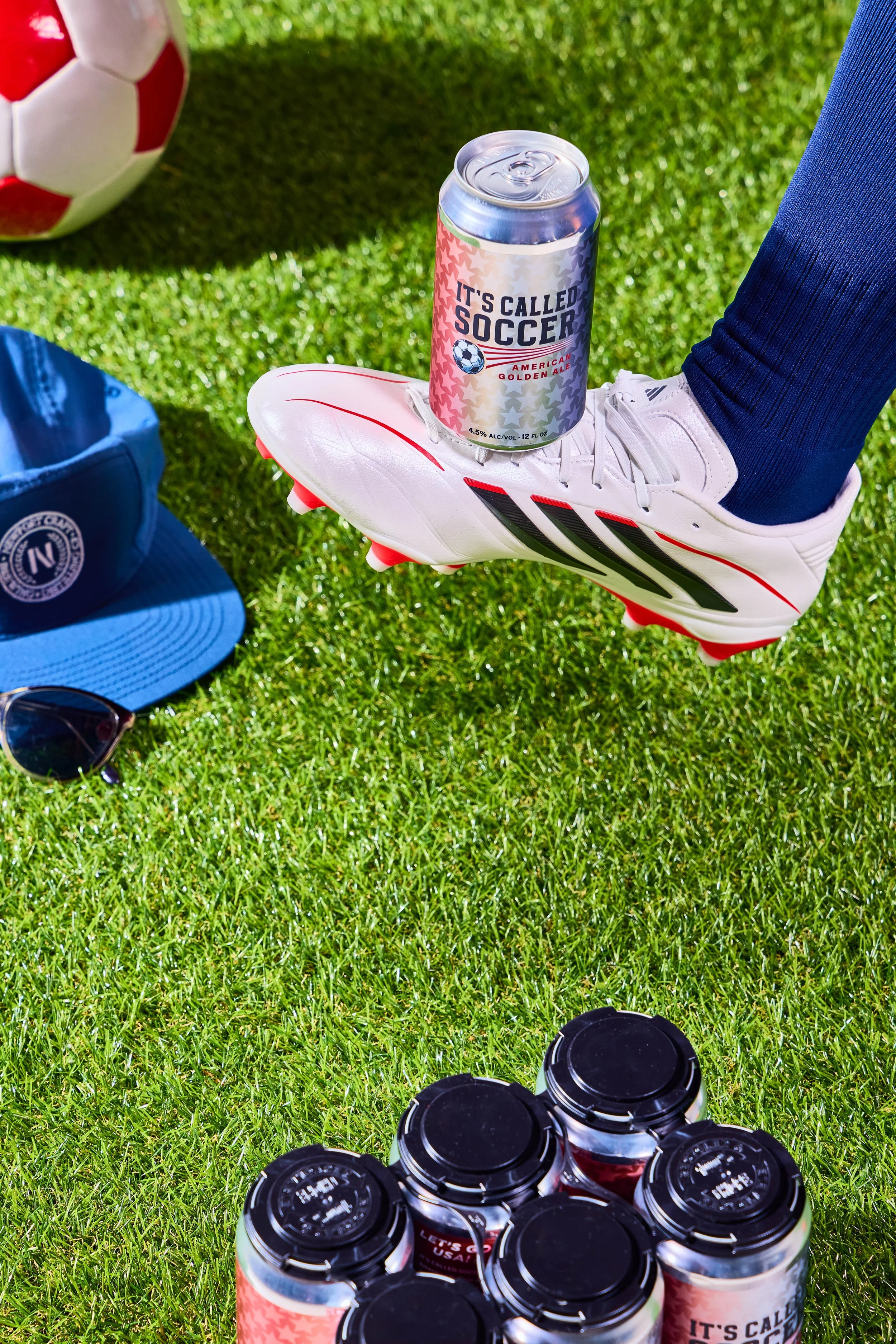 Soccer cleats balanced on a person's foot, a can of beer labeled 'It's Called Soccer,' a soccer ball, a blue baseball cap, sunglasses, and multiple cans of beer on green grass.