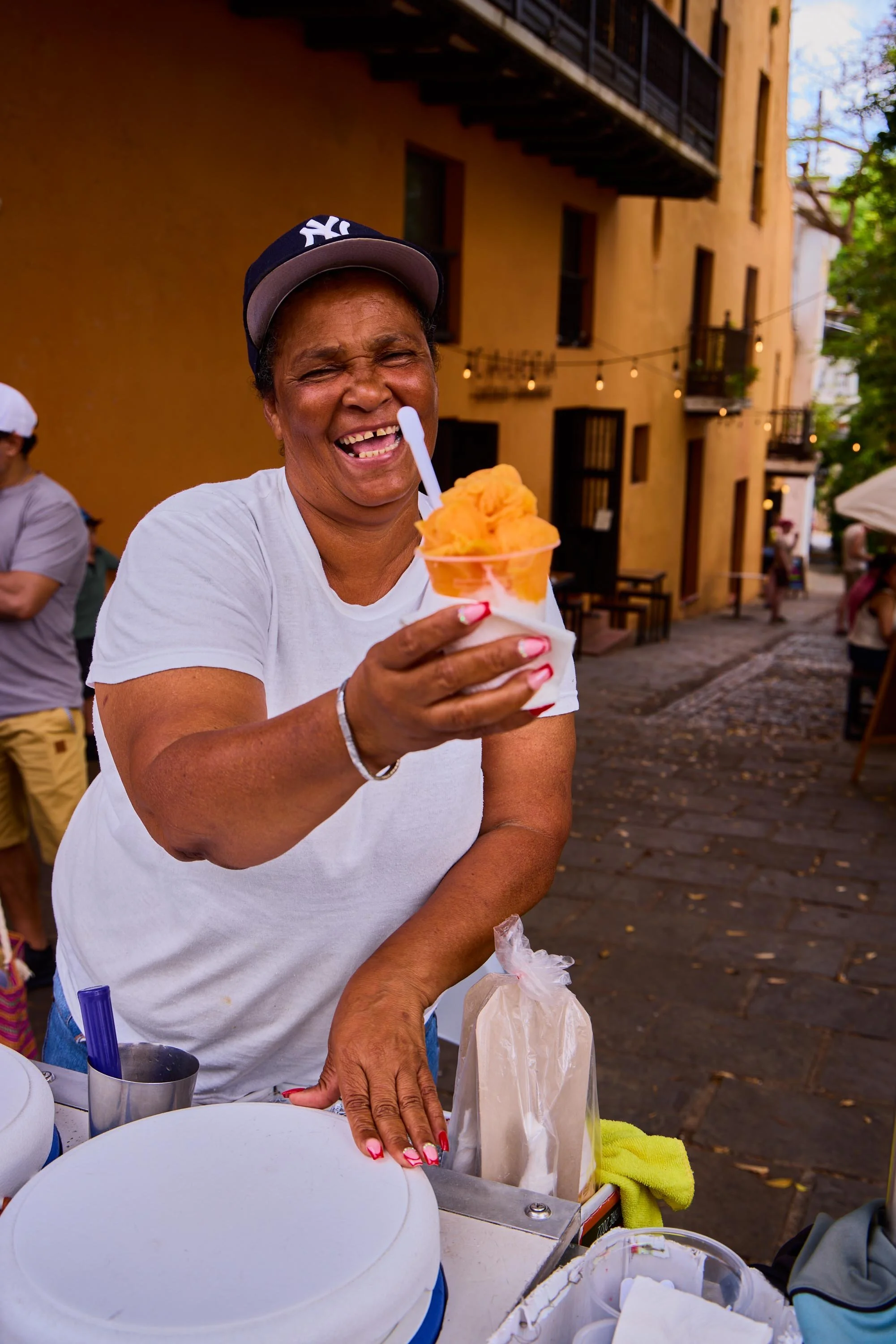 A woman smiling and laughing while holding a cup of bright orange sorbet or ice cream, wearing a baseball cap and a white T-shirt, outdoors with a yellow building and string lights in the background.