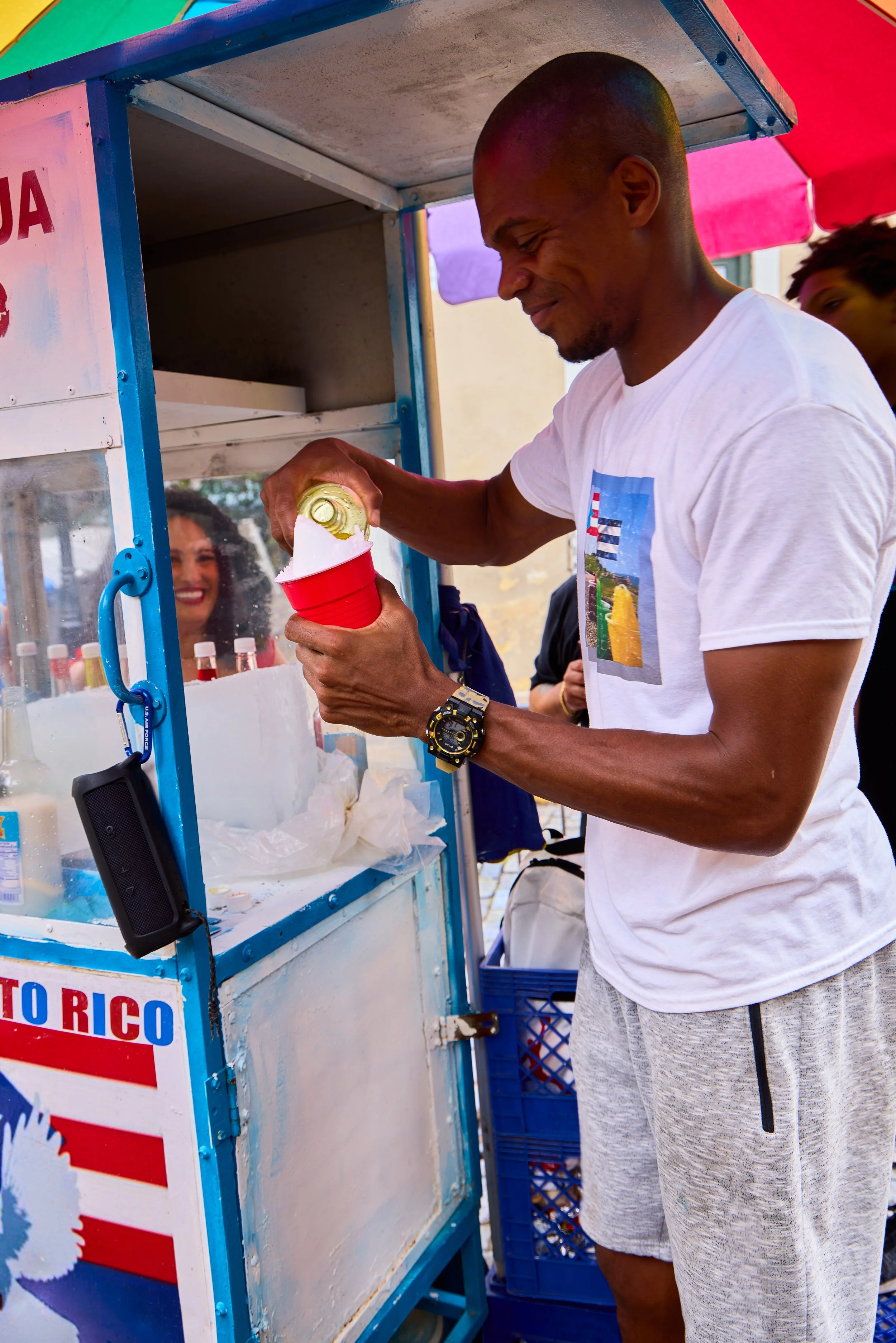 Man pouring a canned drink into a red cup at a colorful street stand.