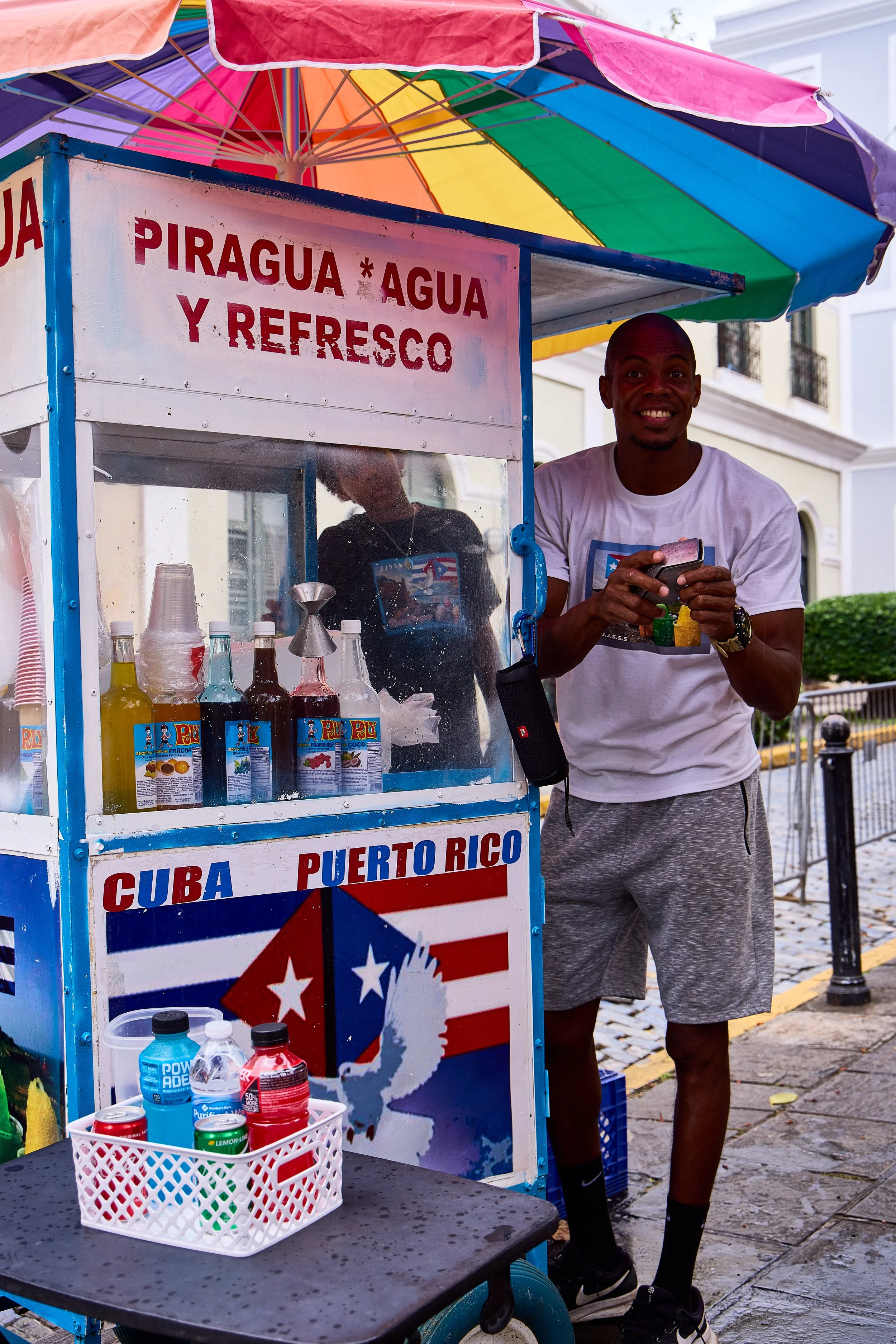 A man standing next to a colorful juice cart under a rainbow umbrella on a sidewalk, with bottles of drinks and a sign mentioning Cuba and Puerto Rico.
