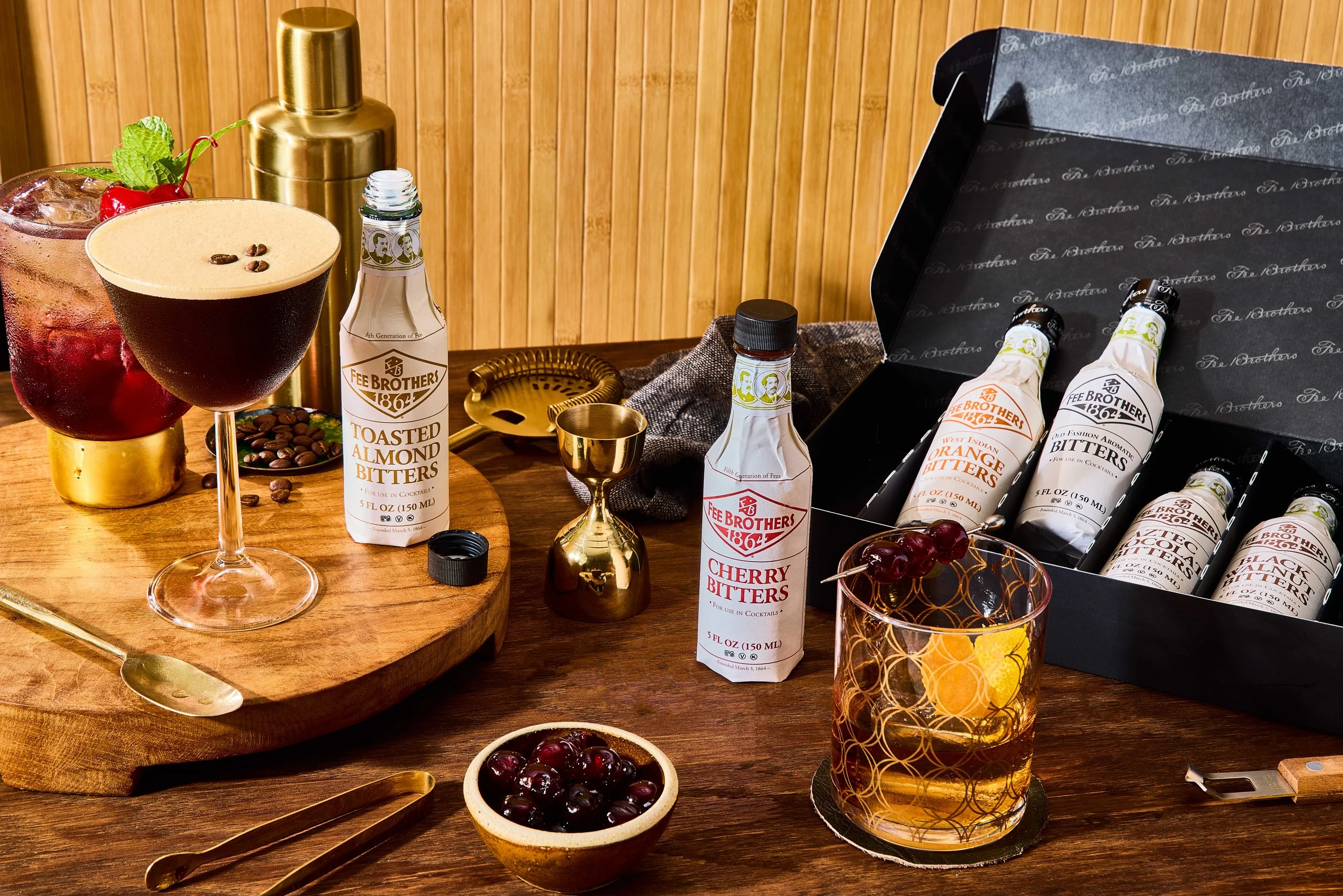 A variety of cocktails and bitters bottles on a wooden table, with cherries, grapes, a gold spoon, a black box of flavored bitters, and bar accessories.