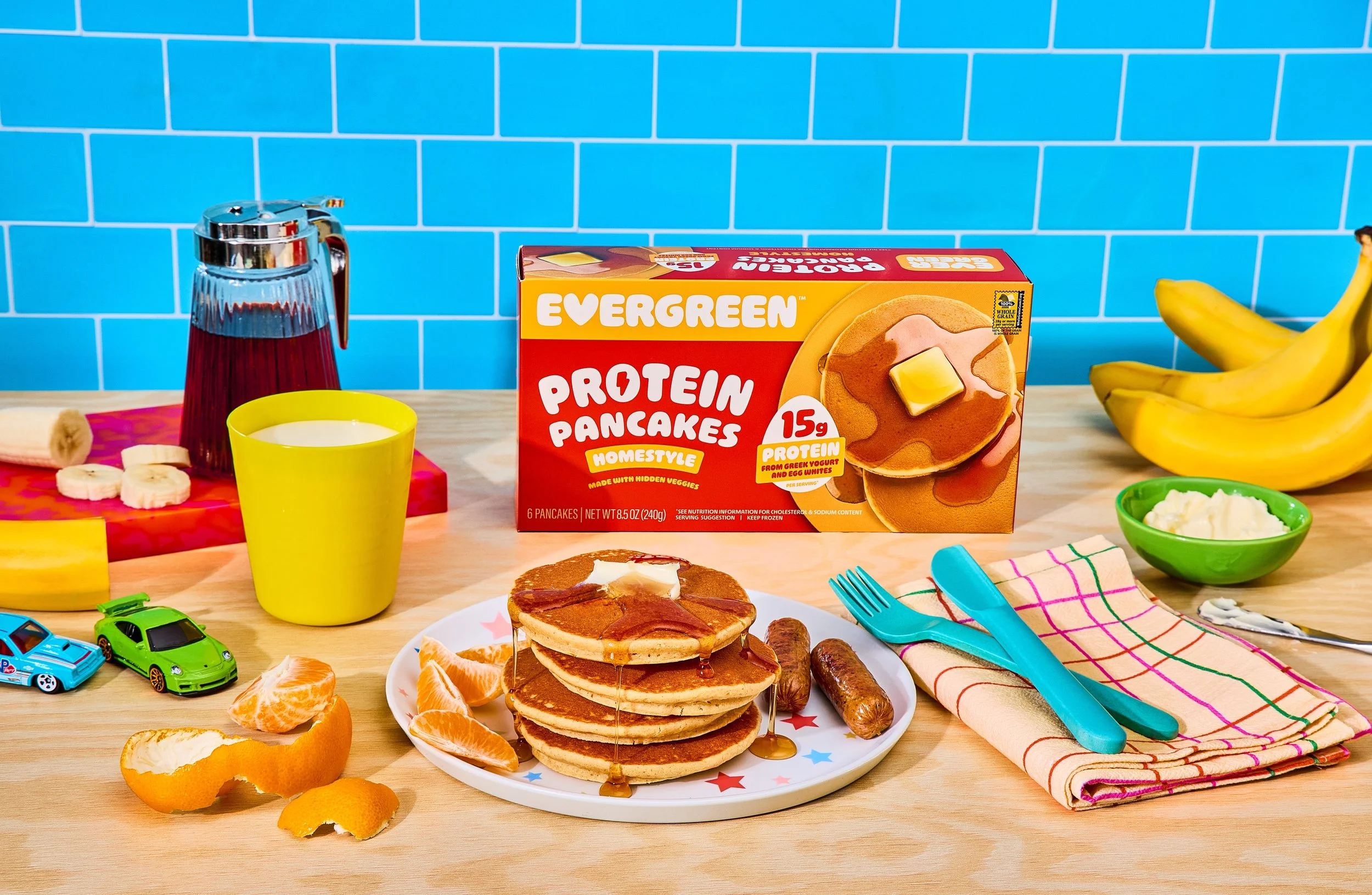 A kitchen or dining table with a box of Evergreen Protein Pancakes, a plate with pancakes topped with syrup and bacon, and various breakfast items including orange slices, peeled orange, and toppings. There are colorful utensils, a yellow cup, a gree