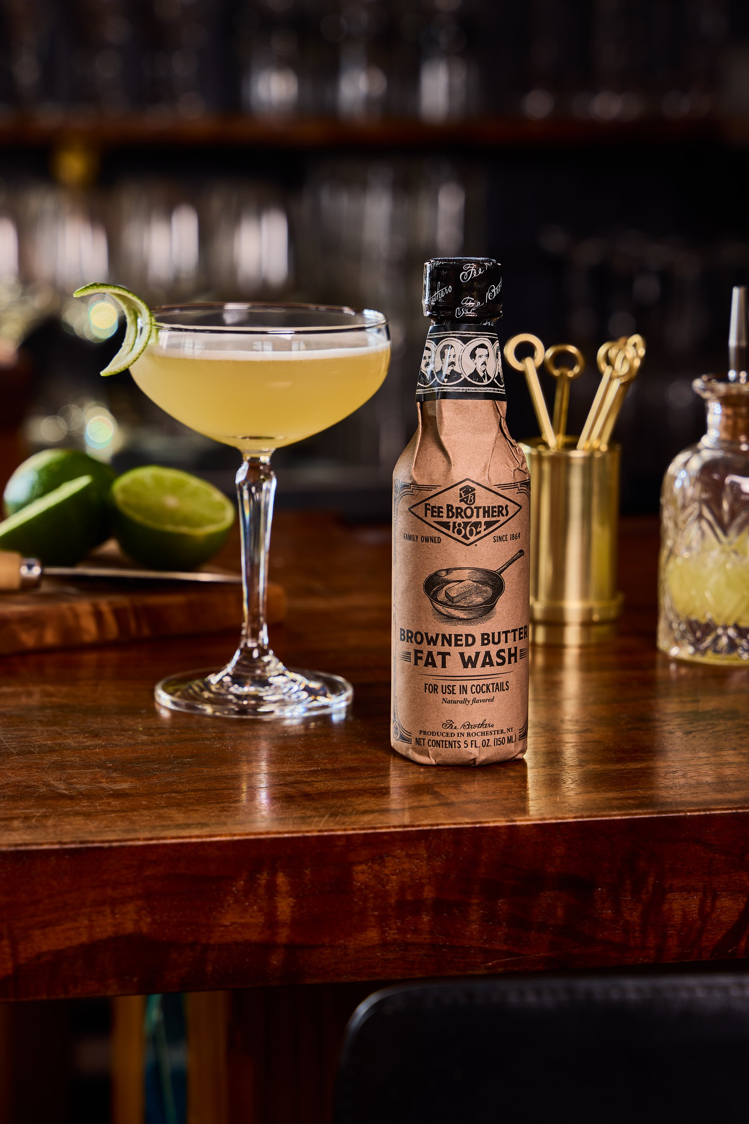 A cocktail glass with a lemon twist garnish, a bottle of Fee Brothers Browned Butter Fat Wash, limes, and bar tools on a wooden bar counter.