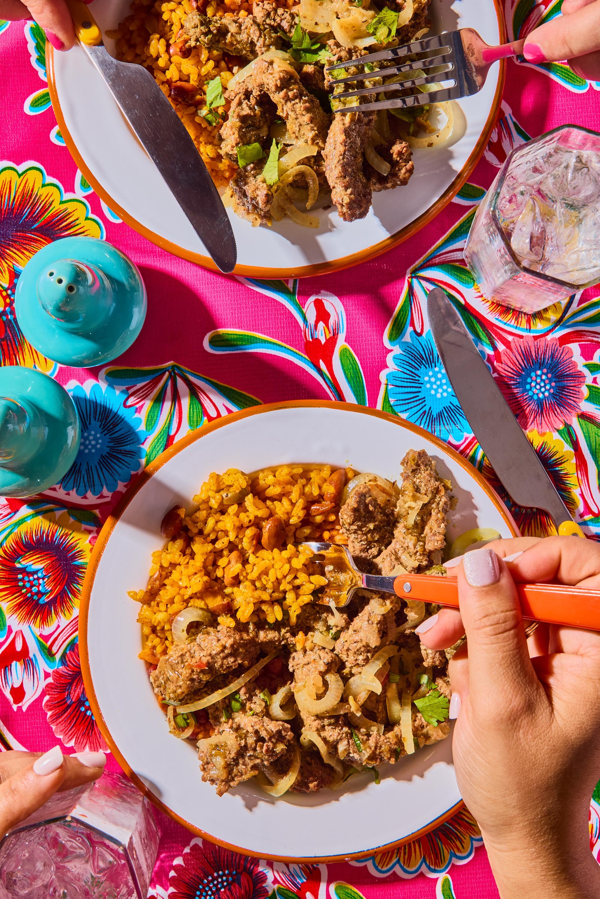 A colorful Mexican meal on a vibrant floral tablecloth, including a plate of seasoned rice, meat with onions, and garnished with cilantro. A person's hand with white nail polish is holding a fork and cutting into the food. There is a glass of ice wat