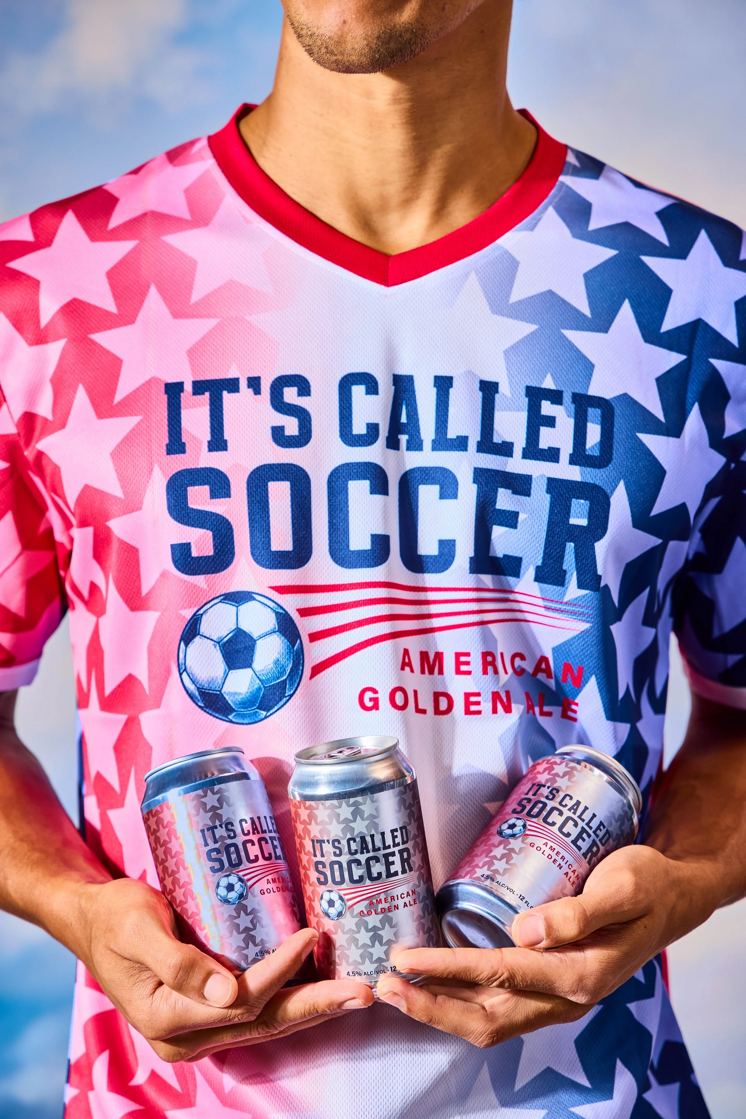 A person wearing a patriotic soccer jersey, holding three cans of American golden ale with a soccer-themed label.