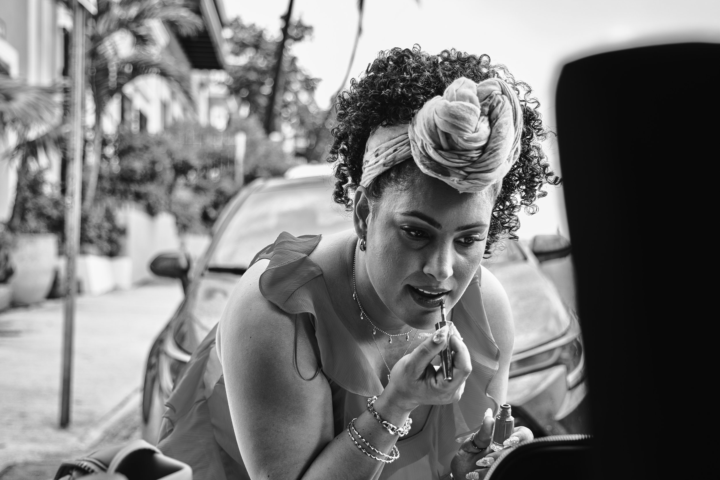 A woman with curly hair wearing a headwrap, jewelry, and a sleeveless top gets her makeup done outdoors. There is a car and some trees in the background.