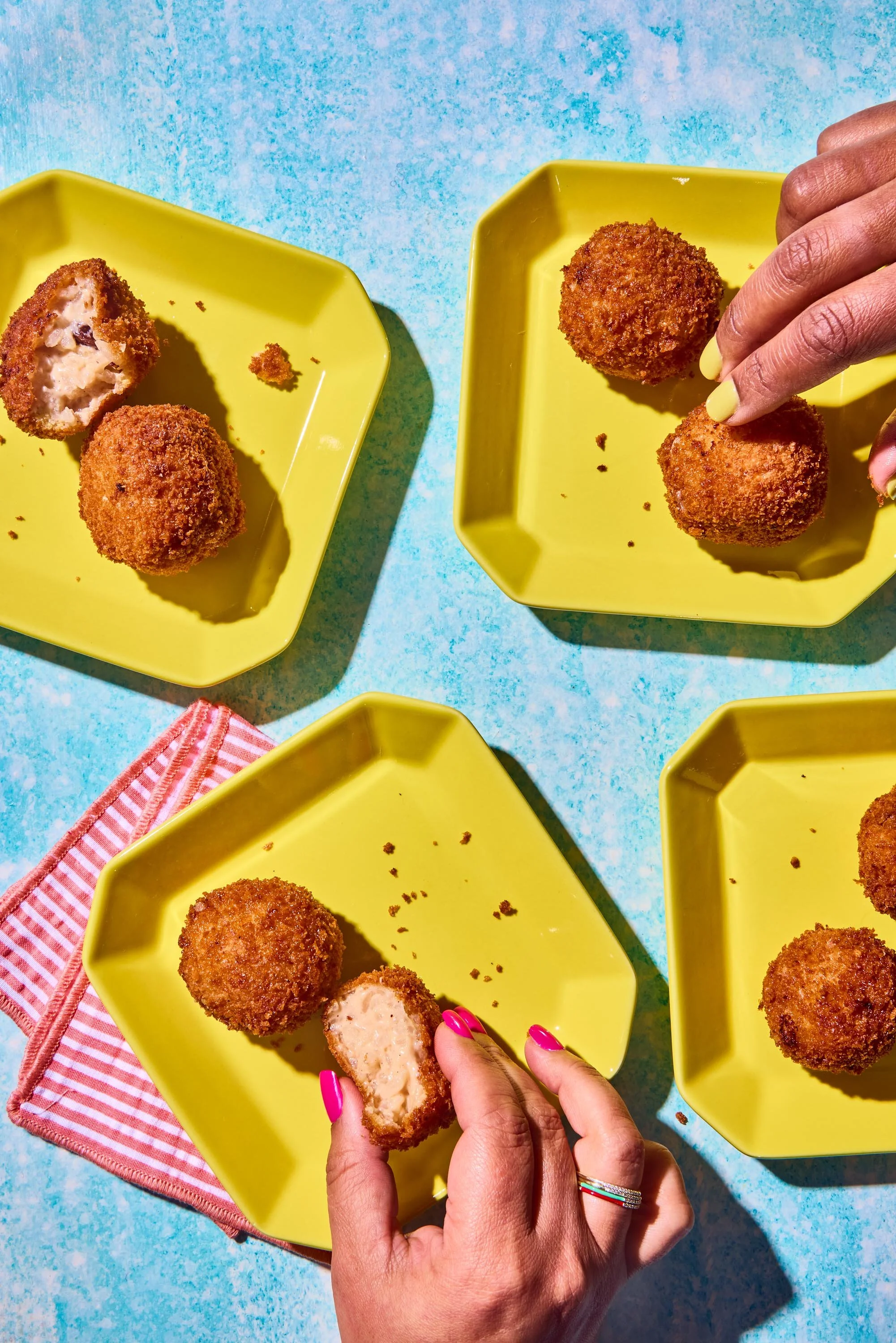 Close-up of person with bright pink nails holding a breaded, fried ball with a creamy filling, on yellow plates with more fried balls on bright yellow plates on a blue surface.