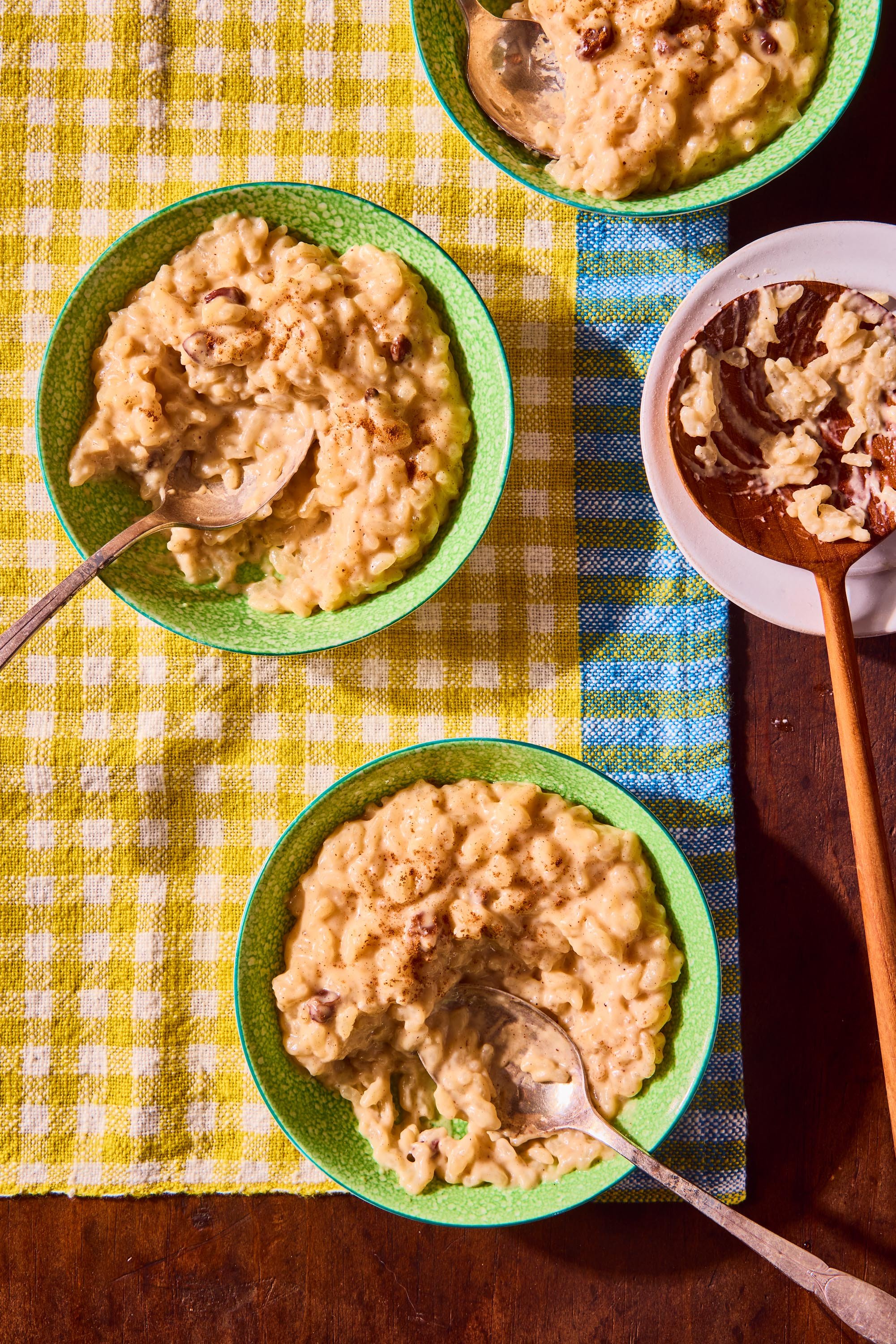 Four bowls of creamy rice pudding with raisins on a table with a yellow checkered cloth and a blue striped cloth underneath, with spoons in each bowl.