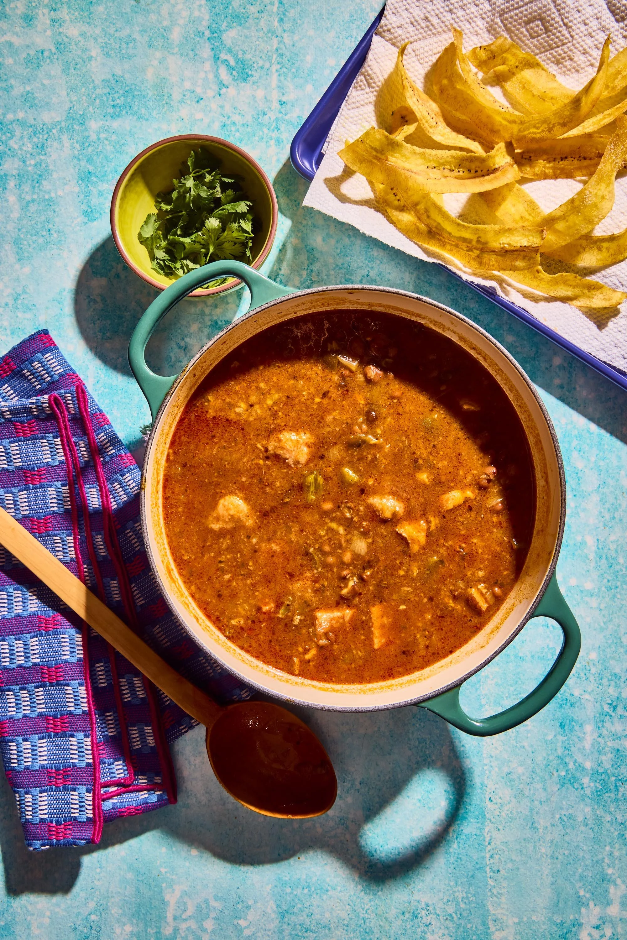 A colorful table setting with a pot of chili, a small bowl of cilantro, a blue tray of banana chips on white paper towels, a wooden spoon, a patterned blue and pink cloth napkin, and a red and pink striped cloth on a blue textured surface.
