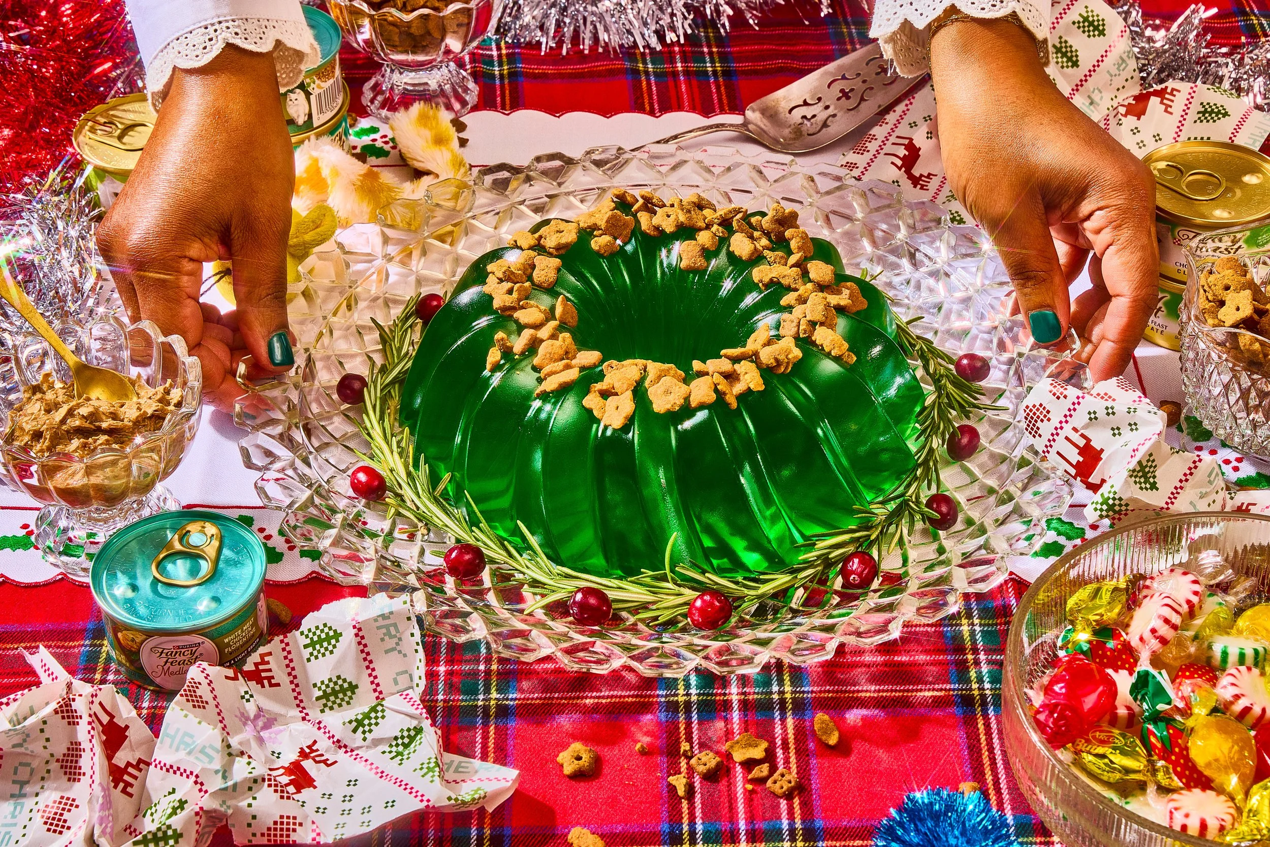 Holiday table with a green gelatin mold topped with animal crackers, surrounded by Christmas treats, candies, canned food, and decorative linens.