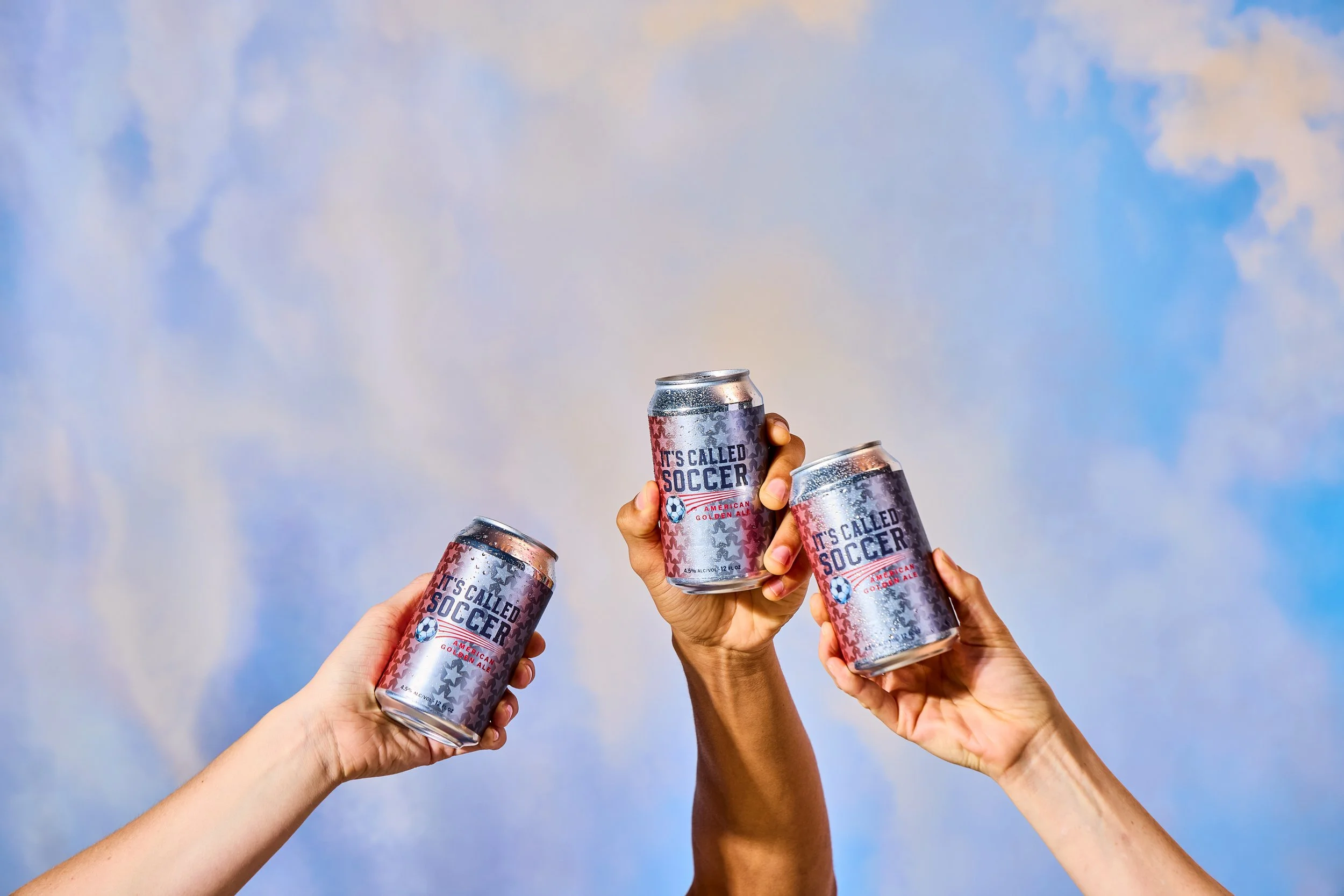 Three hands holding open cans of soda with the label 'It's Called Soccer' against a cloudy sky background.
