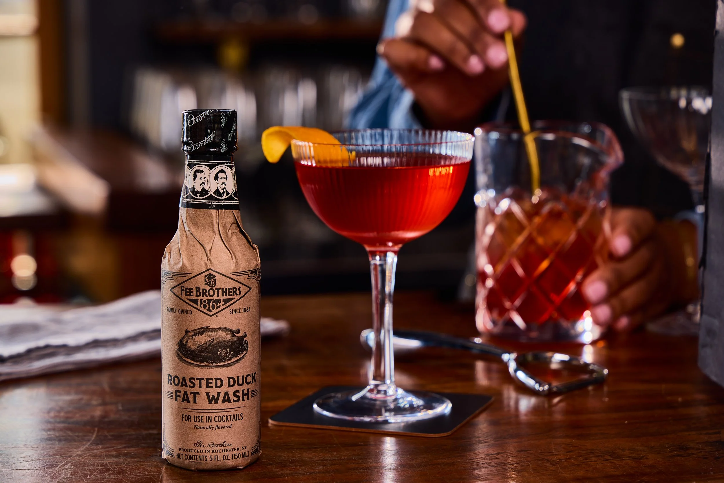 A bottle of Fee Brothers roasted duck fat wash and a cocktail glass with a red drink garnished with a lemon peel on a bar counter. In the background, a person's hand is stirring another drink in a glass tumbler.