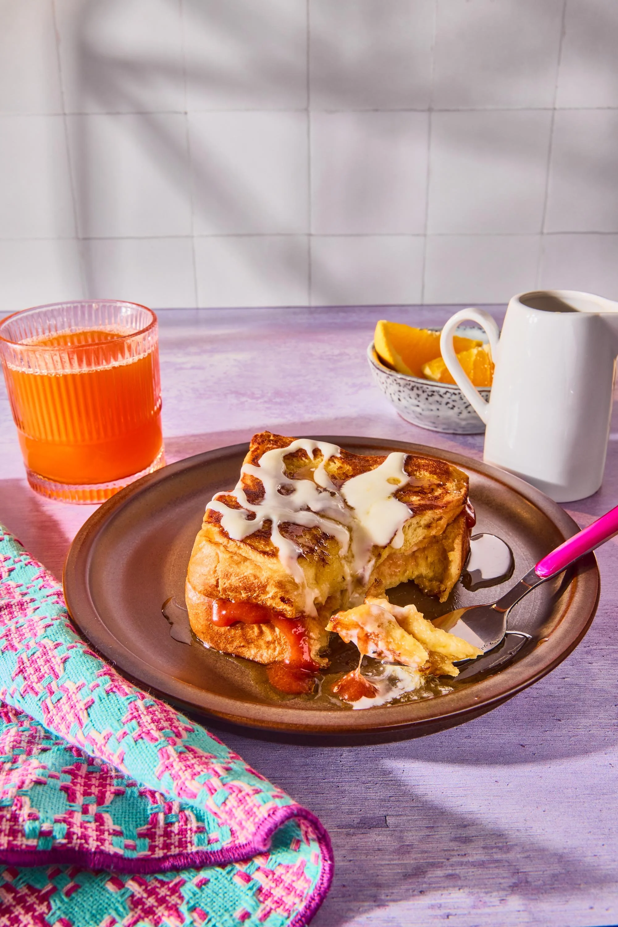 A breakfast plate with a slice of French toast topped with white sauce on a brown plate. Beside it, a fork is partially inserted. In the background, a glass of orange juice, a small bowl of orange slices, a white mug with a dark beverage, and a pink 