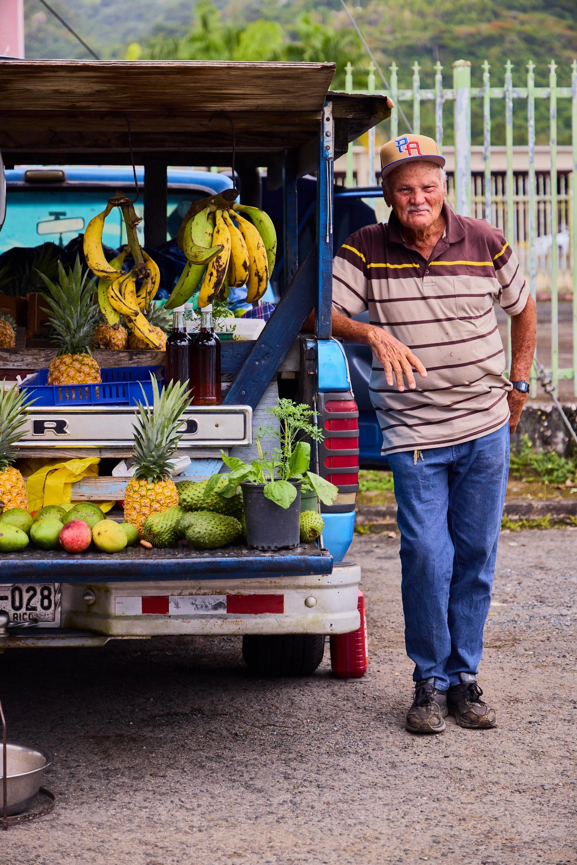 An elderly man stands beside a small fruit stand attached to a pickup truck, with bananas, pineapples, and other fruits on display, in an outdoor market with a green, hilly background.