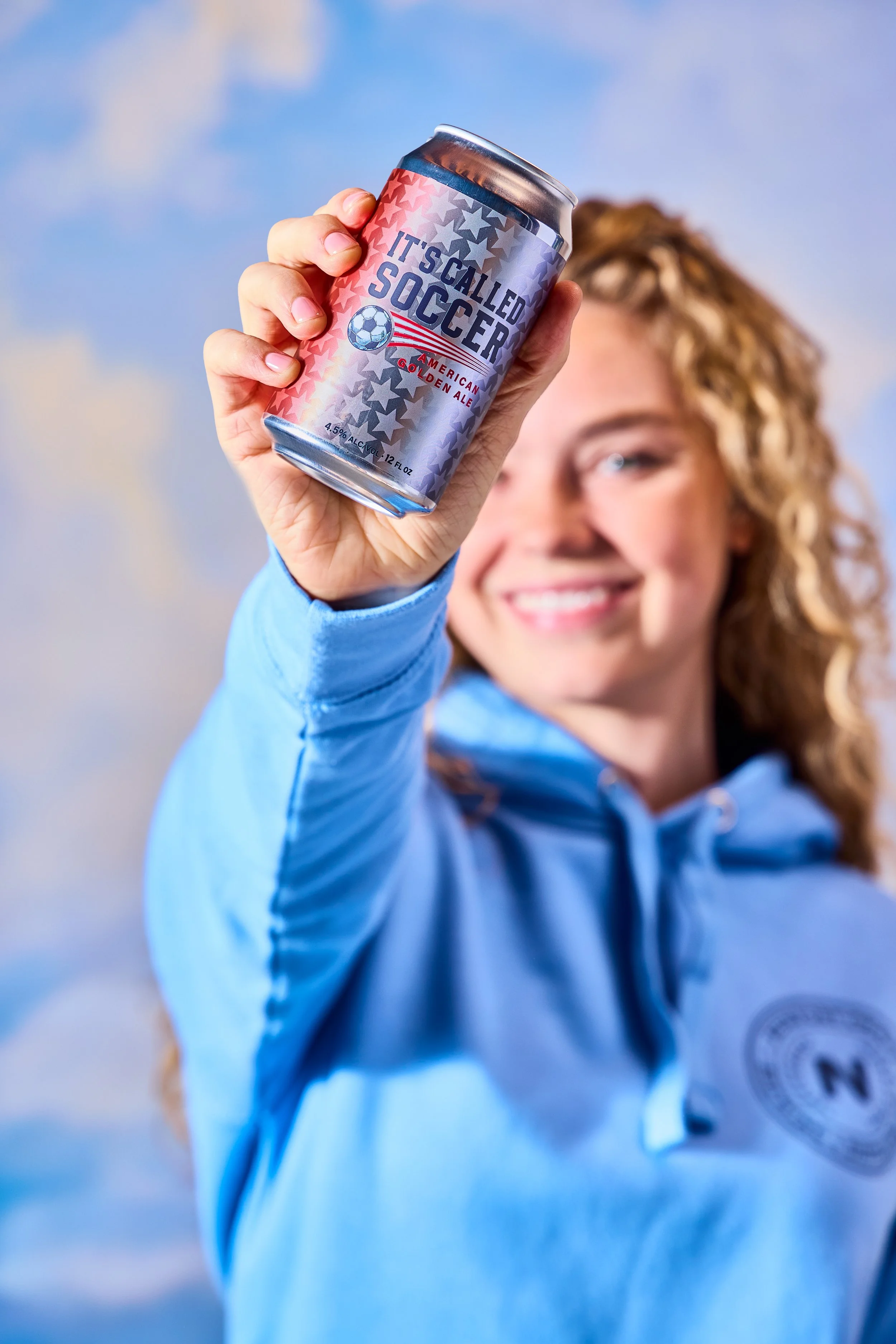 Young woman with blonde curly hair smiling and holding a soda can labeled 'It's Called Soccer' in front of her, with a partly cloudy blue sky in the background.