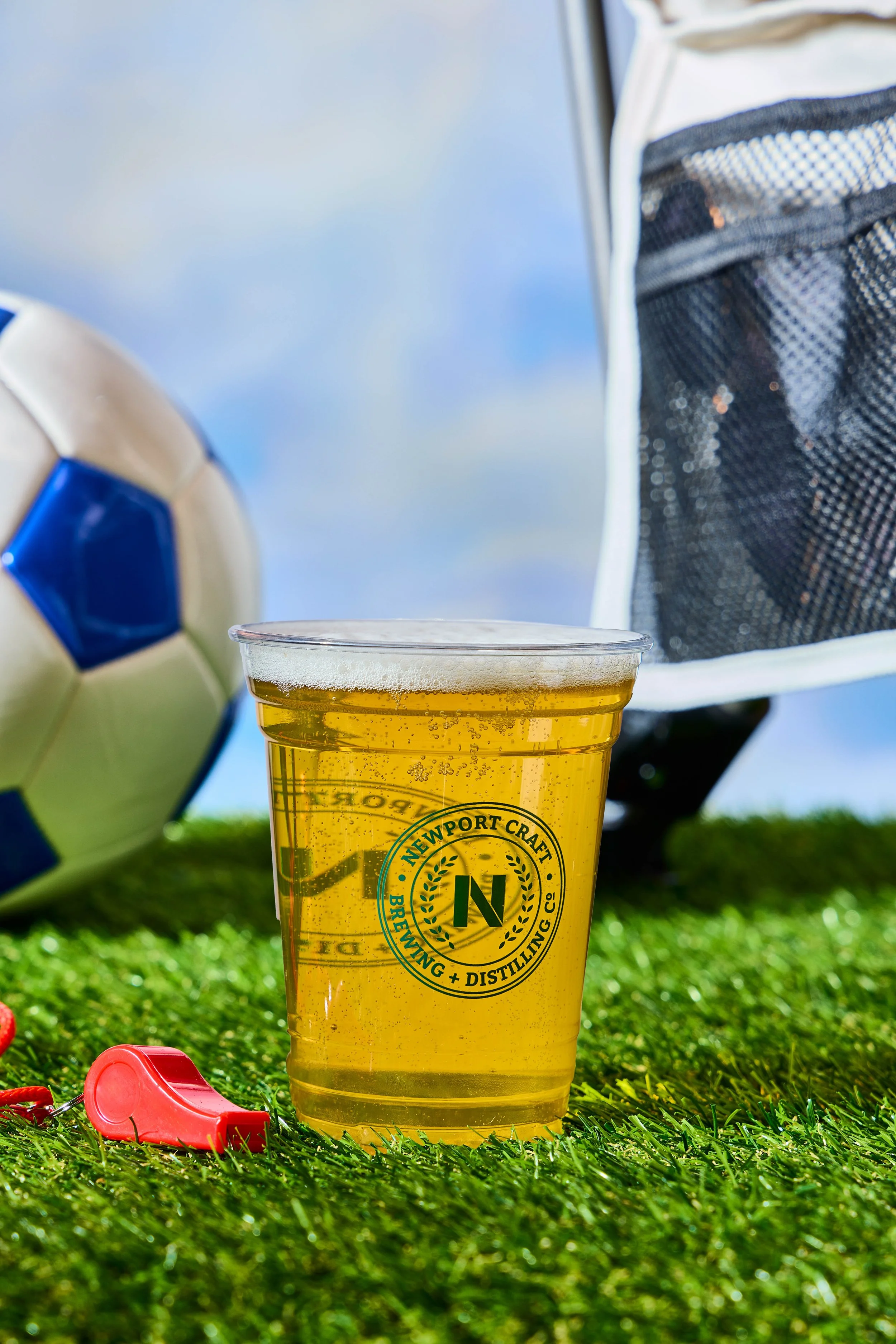 A clear plastic cup filled with a golden beer with bubbles, placed on artificial green grass. A soccer ball is partially visible on the left, and a black and white soccer net is in the background on the right.