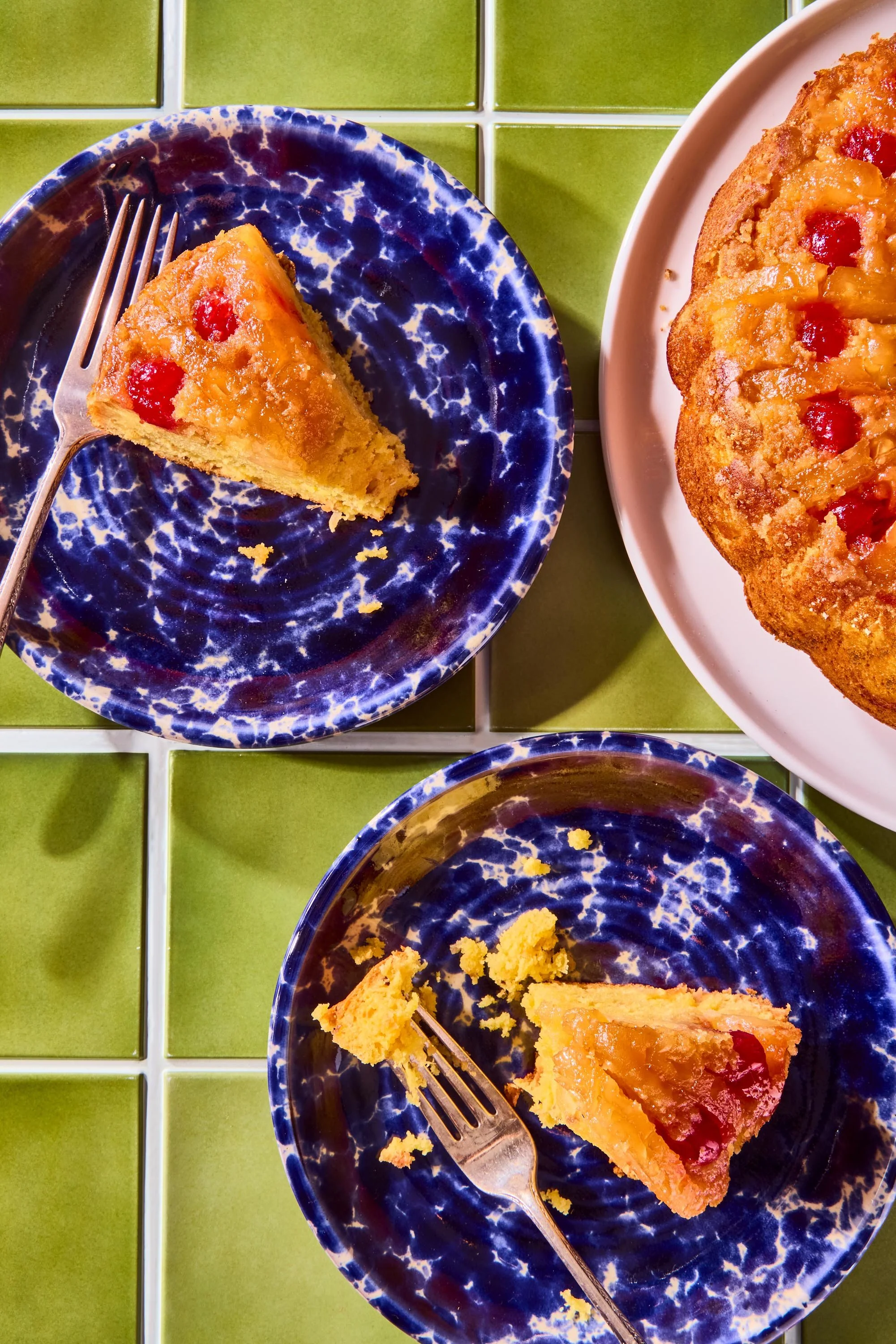 A slice of slice pie with cherries and pineapple on a blue patterned plate, a fork, and a whole pie with cherries and pineapple on a white plate, all on a green tiled surface.