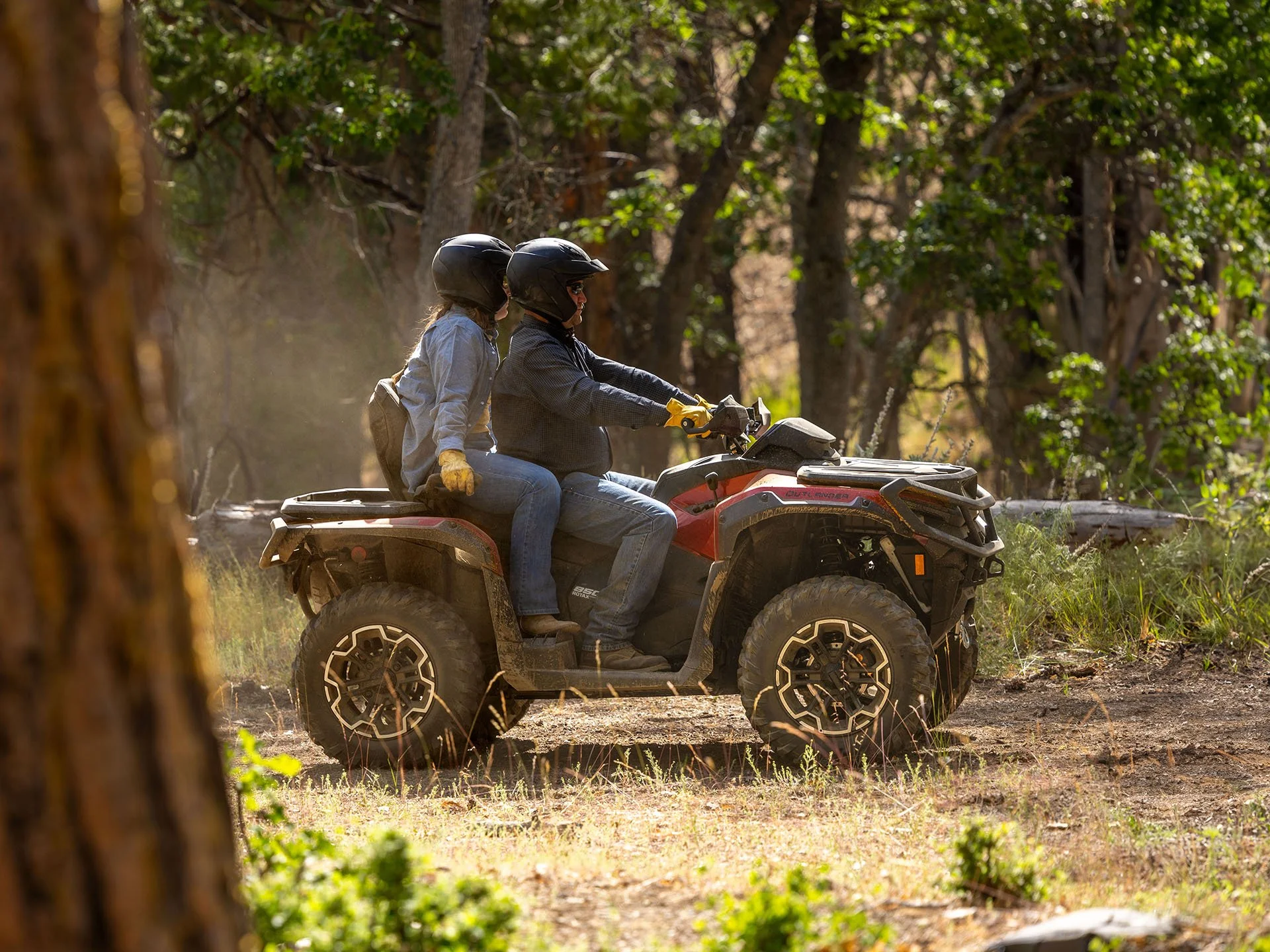 A man and a woman riding a red all-terrain vehicle through a wooded area, both wearing helmets, with tall trees and green foliage in the background.