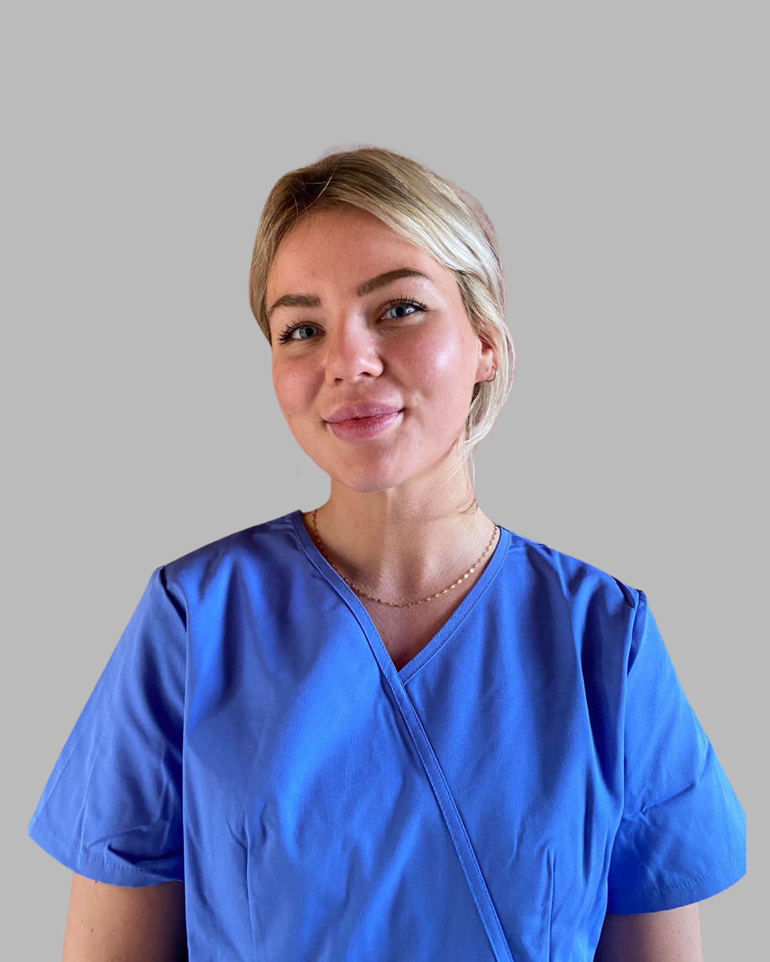 A woman with blonde hair wearing a blue medical scrub top and a delicate gold necklace, smiling softly at the camera against a plain background.