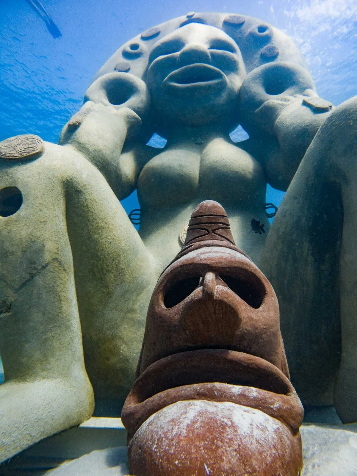 Underwater photo of a large stone sculpture of a giant head with one hand resting on its chin, surrounded by rocks and a fish swimming nearby.