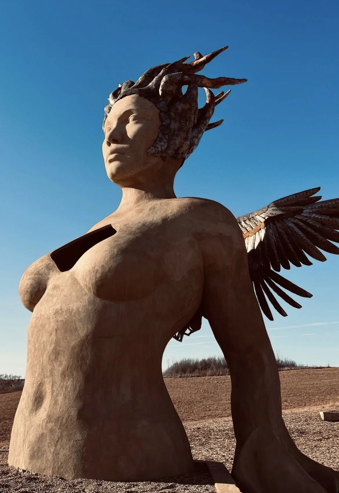 A large sculpture of a mythological woman with wings, a headdress resembling tree branches, and a black open space on her chest, situated in an open field with a person sitting nearby.