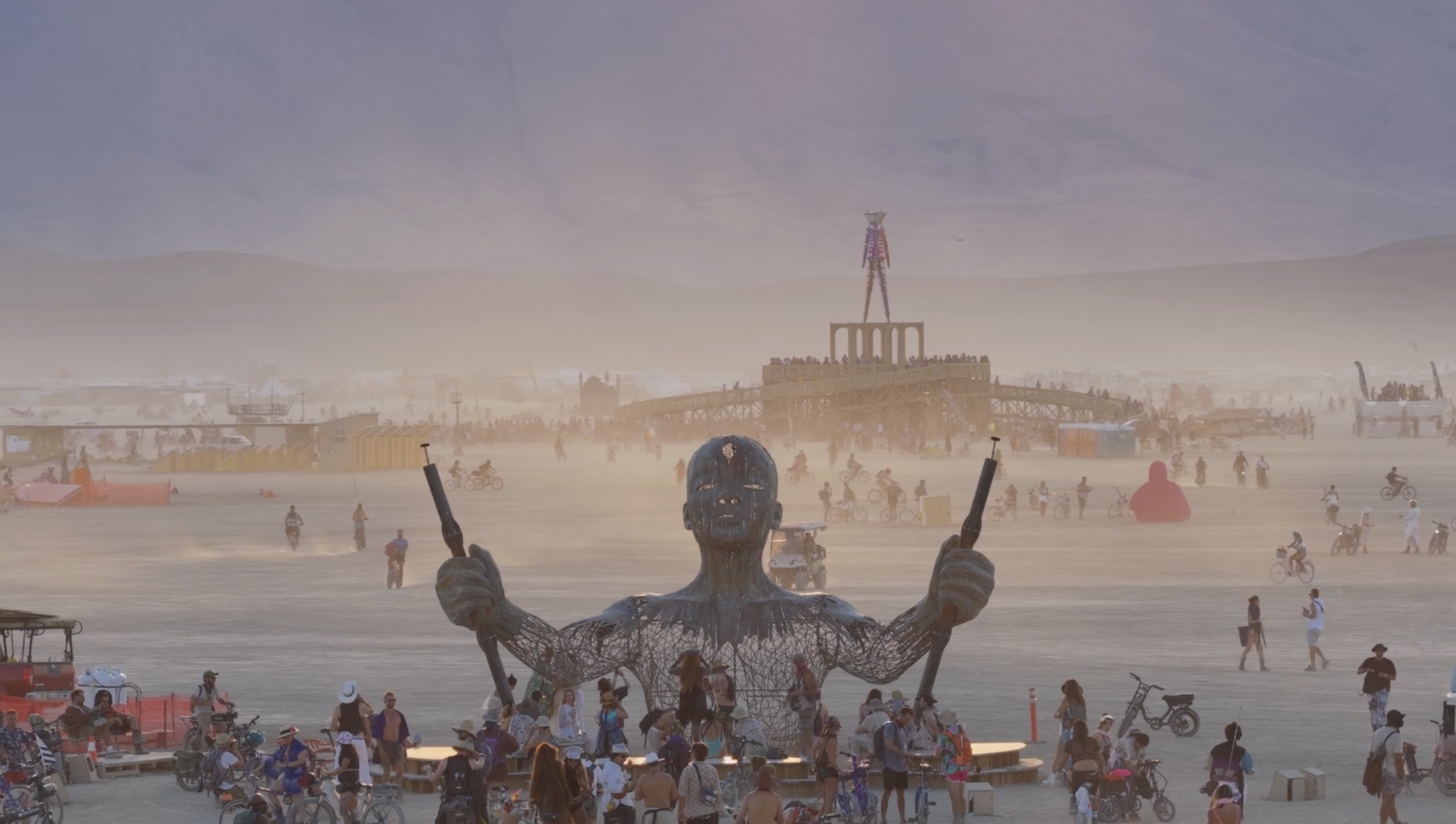 Large art installation of a woman with wooden skin, holding two rifles, with mountains in the background and a desert landscape.