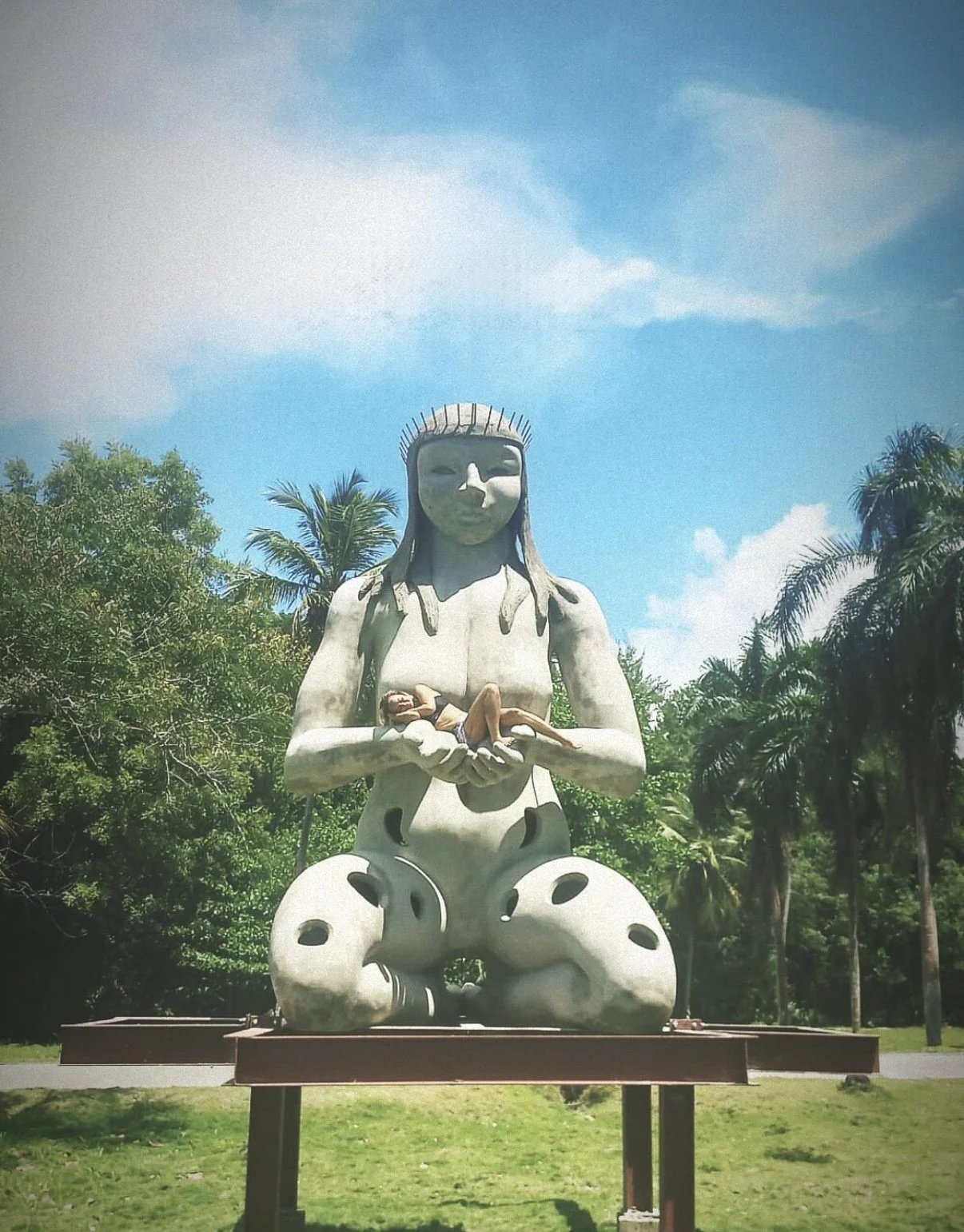 A large outdoor sculpture of a woman with long hair holding a small nude figure, set against a background of green trees and a partly cloudy sky.