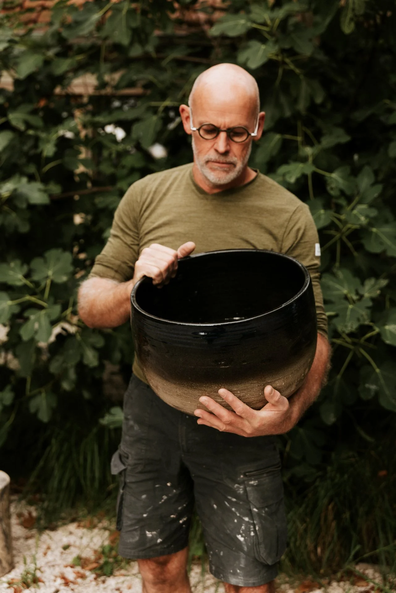 Een man met een kale kop en brillen houdt een groot zwart gebakken pot vast.