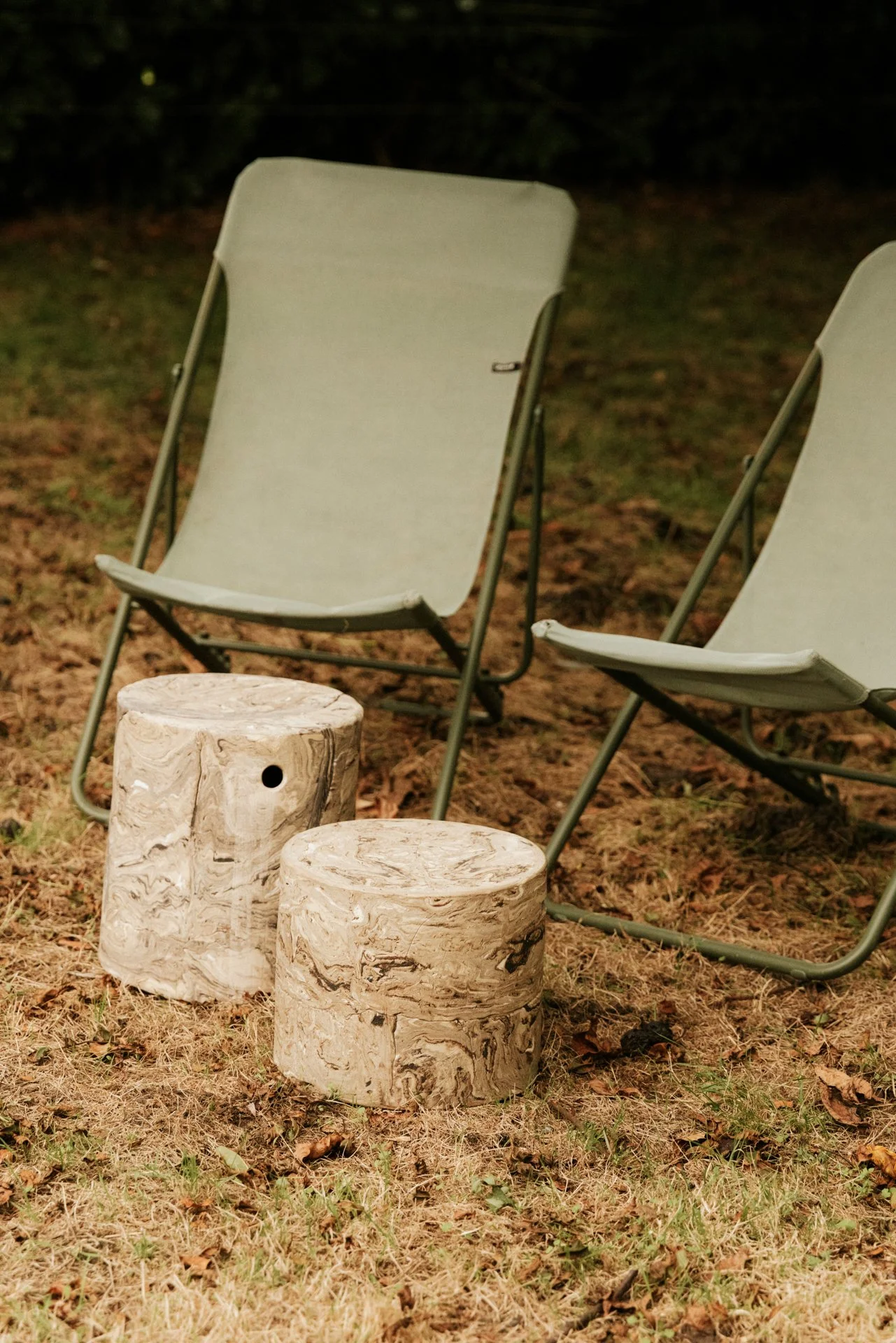Twee beige houten stokjes en twee beige campingstoelen op een grasveld met bladeren, 's avonds of in de schemering.