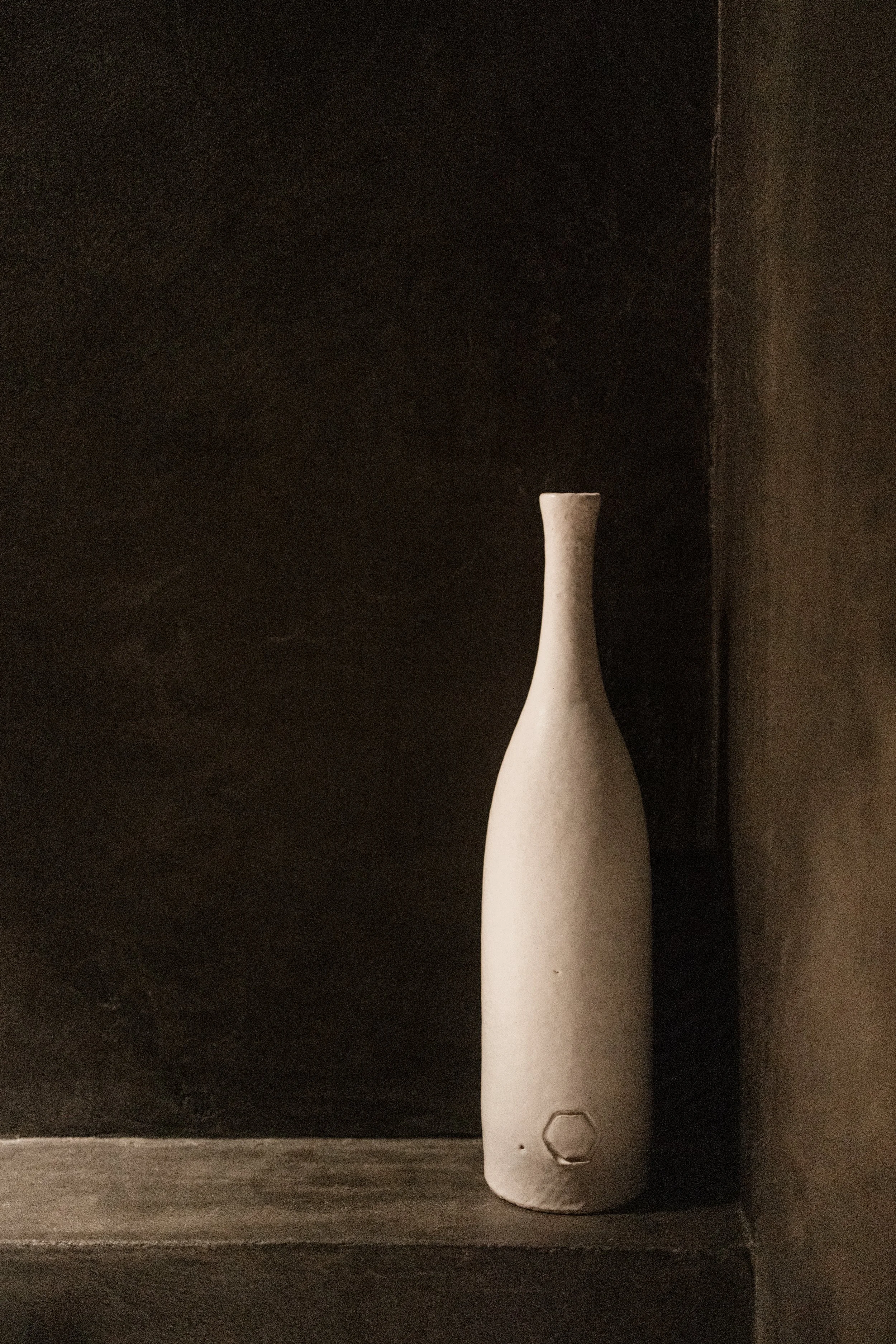Een witte keramieken vaas met een slanke, gebogen vorm, geplaatst op een donkere houten plank tegen een donkerbruine muur.