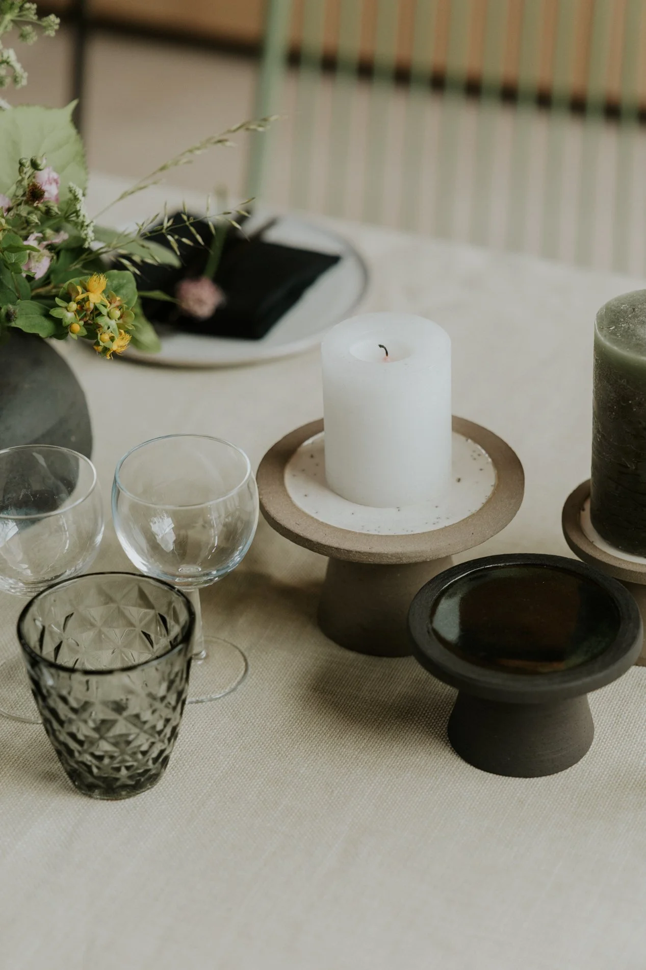 Een tafel met een witte kaars, glazen en een bloemenmand, op een beige tafelkleed.