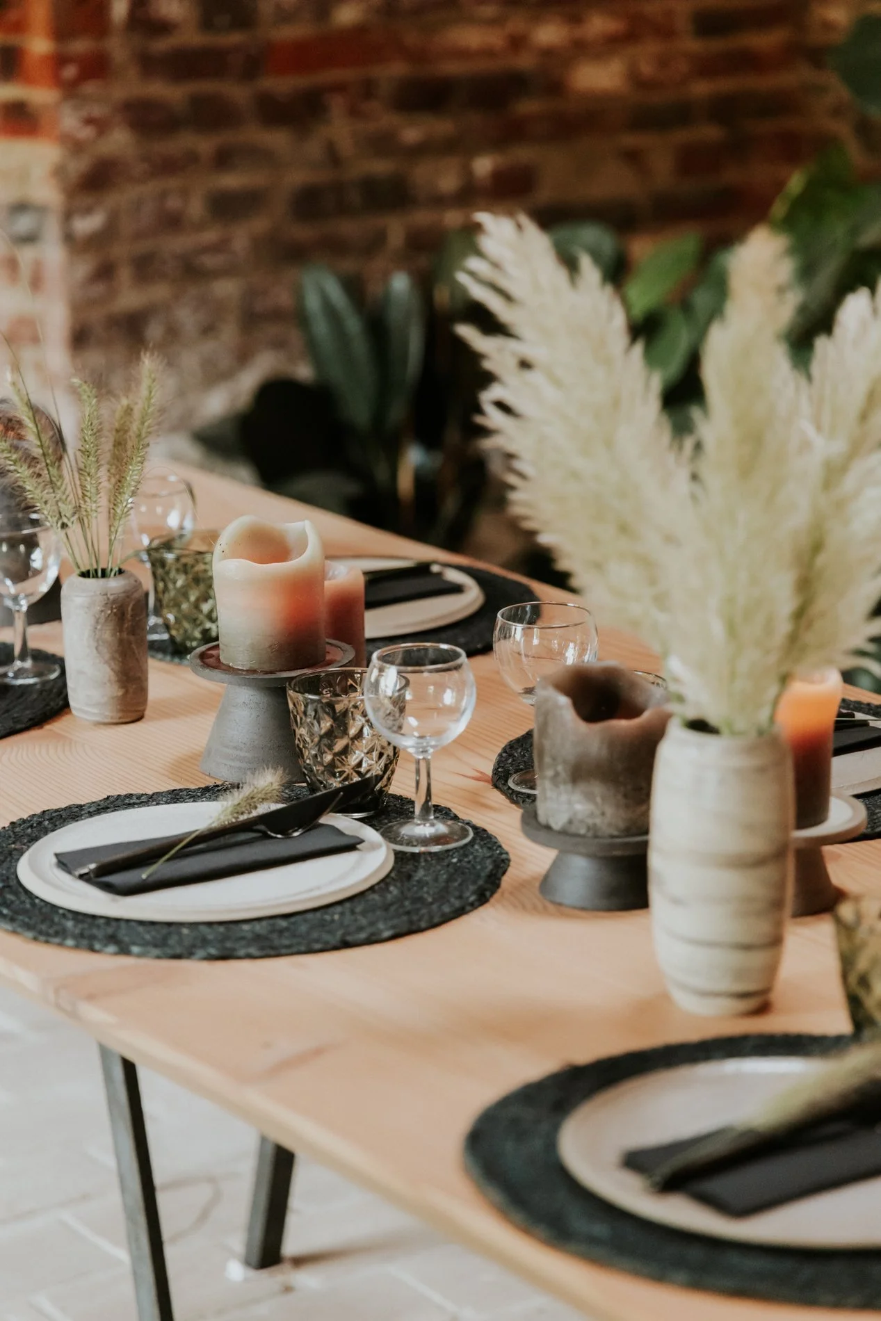 Een feesttafel met gedekte borden, glazen en kaarsen, versierd met vazen met gedroogde bloemen en pampasgras, tegen een bakstenen muur.