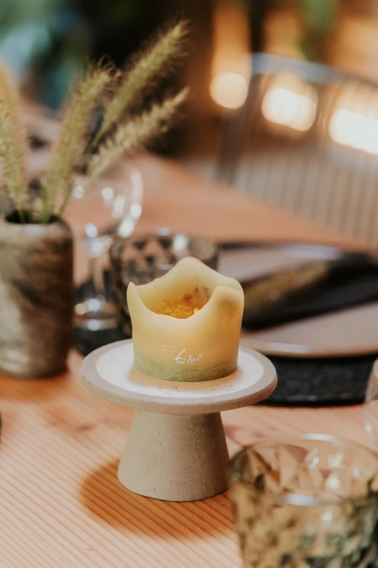 Een keramieken kandelaar met een witte kaars op een houten tafel, met op de achtergrond een vaas met planten en een boek.