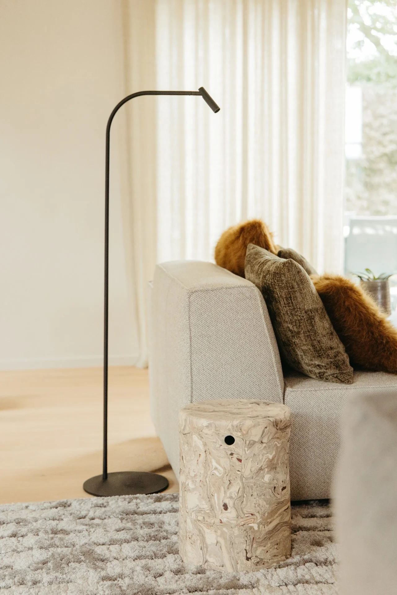 Woonkamer met een lichtgrijze bank, twee verschillende kussens, een houten bijzettafel, een zwarte vloerlamp en een grote raam met witte gordijnen die natuurlijk licht binnenlaten.
