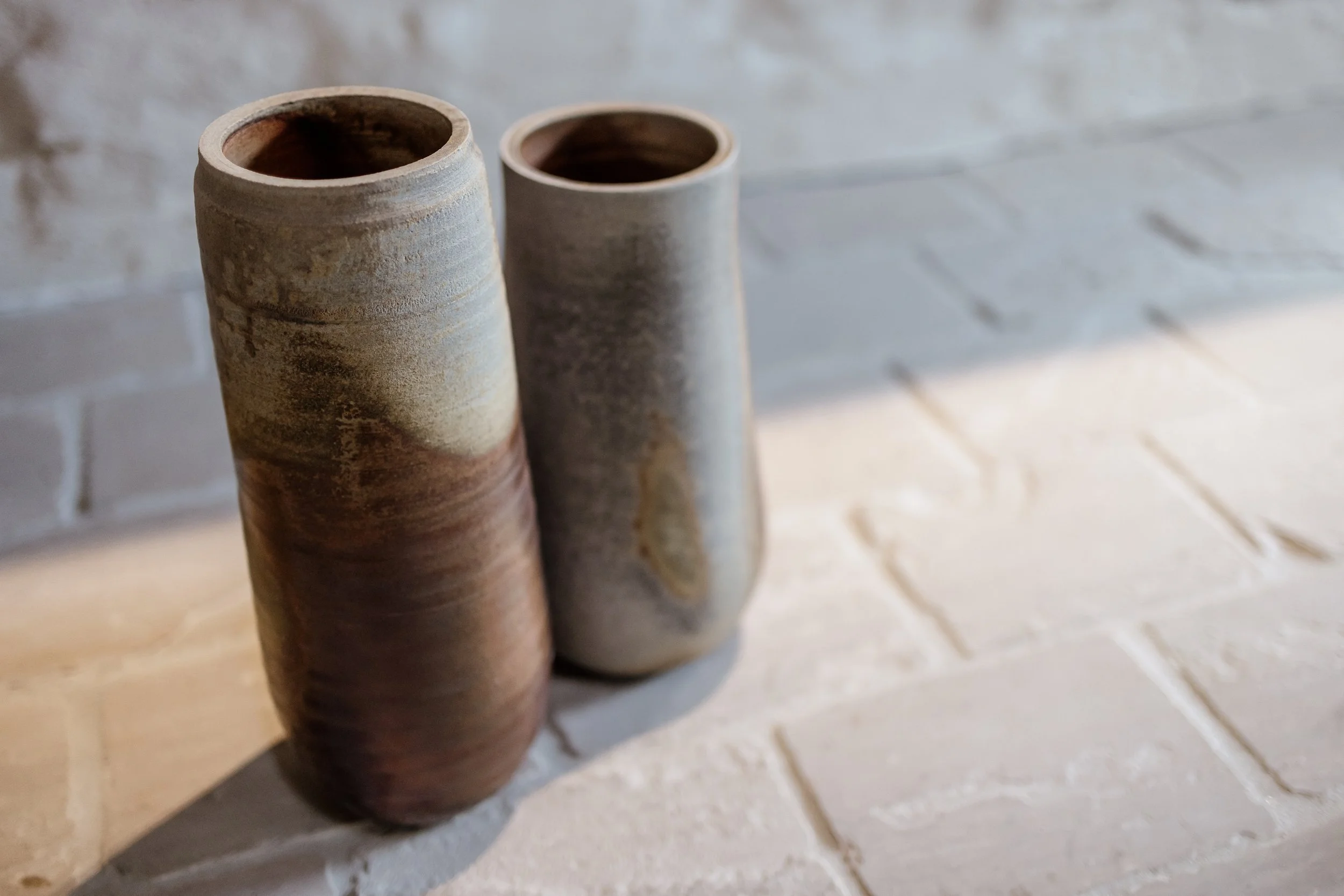 Twee lange, cilindrische, aardewerken vazen met een ruwe, natuurlijke textuur, geplaatst op een lichte, betegelde vloer met een witte bakstenen muur op de achtergrond.
