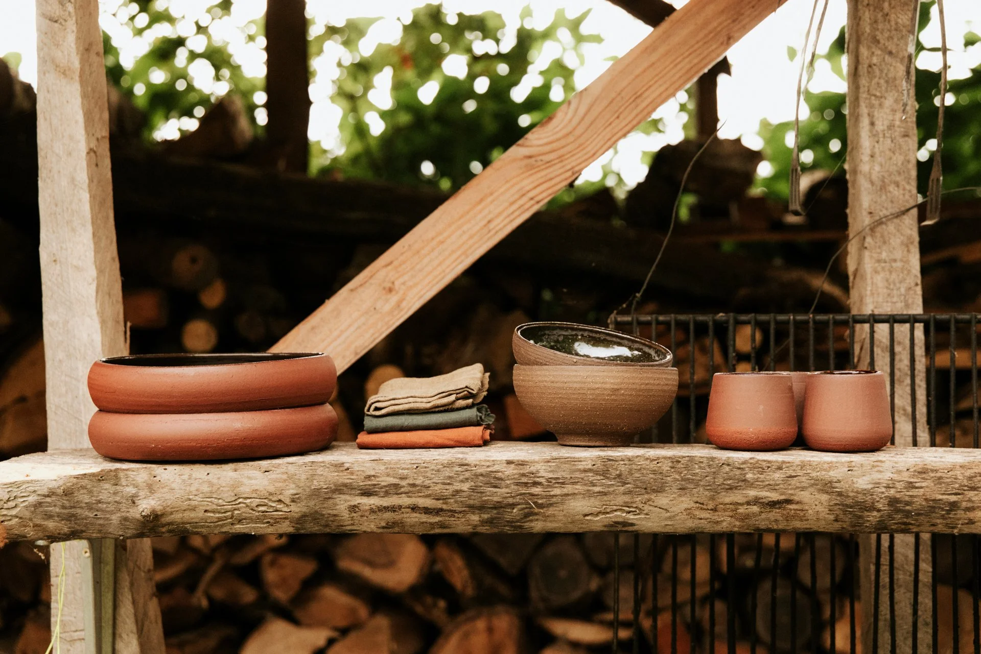 Schalen in keramiek die rood gekleurd zijn op een houten bank met handdoeken er naast, achtergrond tuin en houtberging.