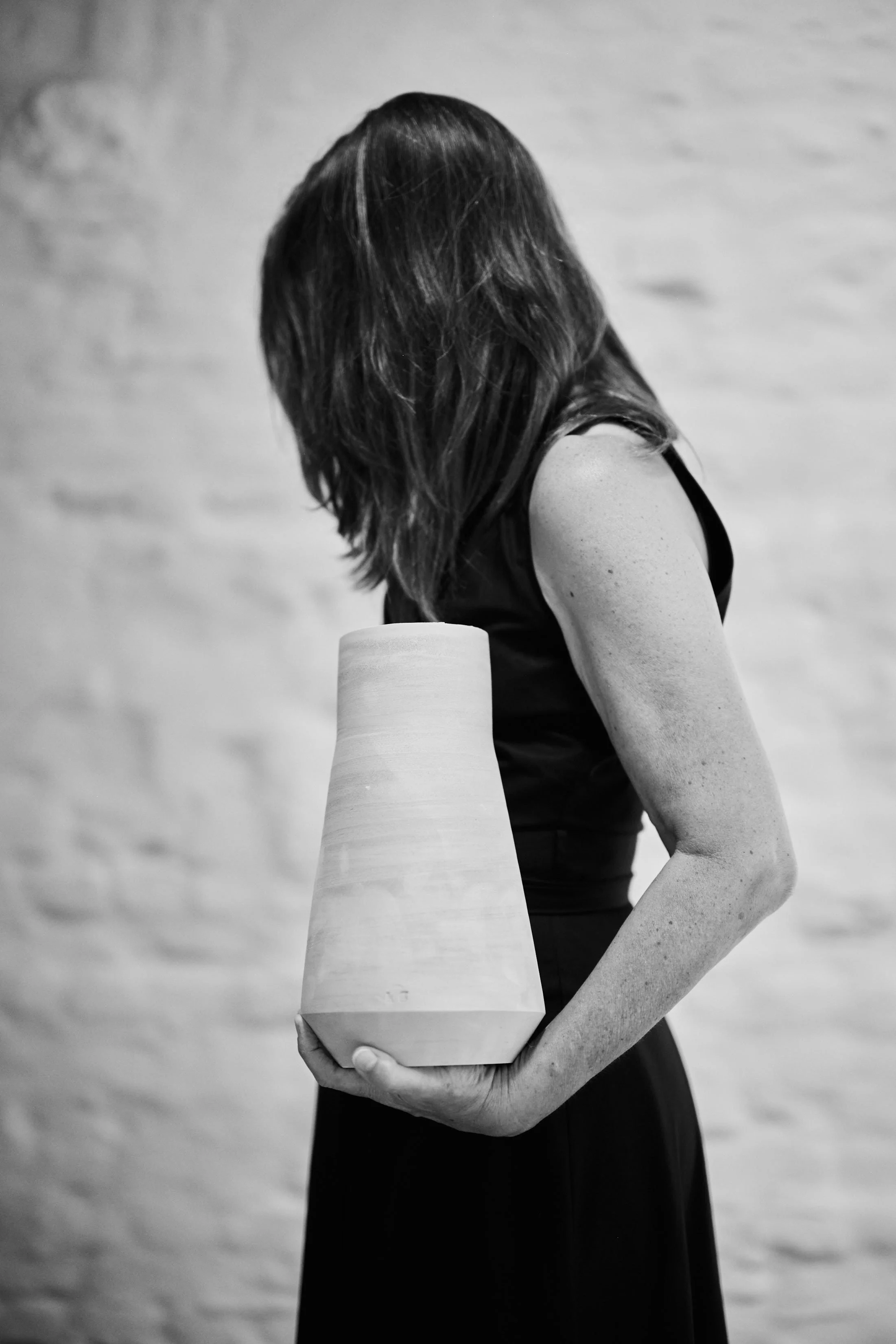Een vrouw die een grote vaas vasthoudt, met lang haar en een zwart jurk, zonder gezicht zichtbaar, op een effen achtergrond.