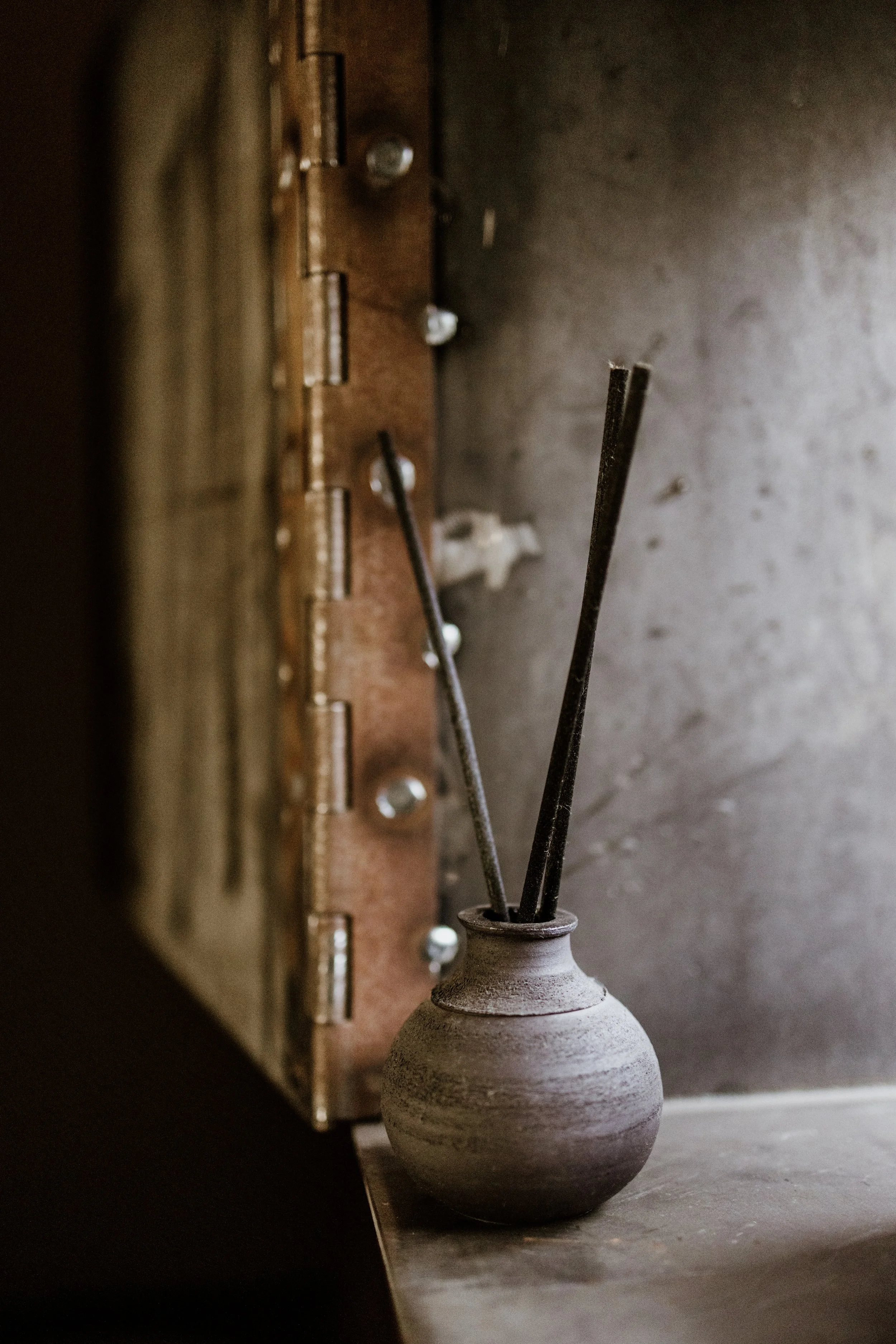 Een grijze keramieken vaas met een smalle hals en een bolvormige onderkant, gevuld met twee zwart geurstokjes, staat op een houten plank. Op de achtergrond zit een houten apparaat met metalen schroeven en bevestigingen.