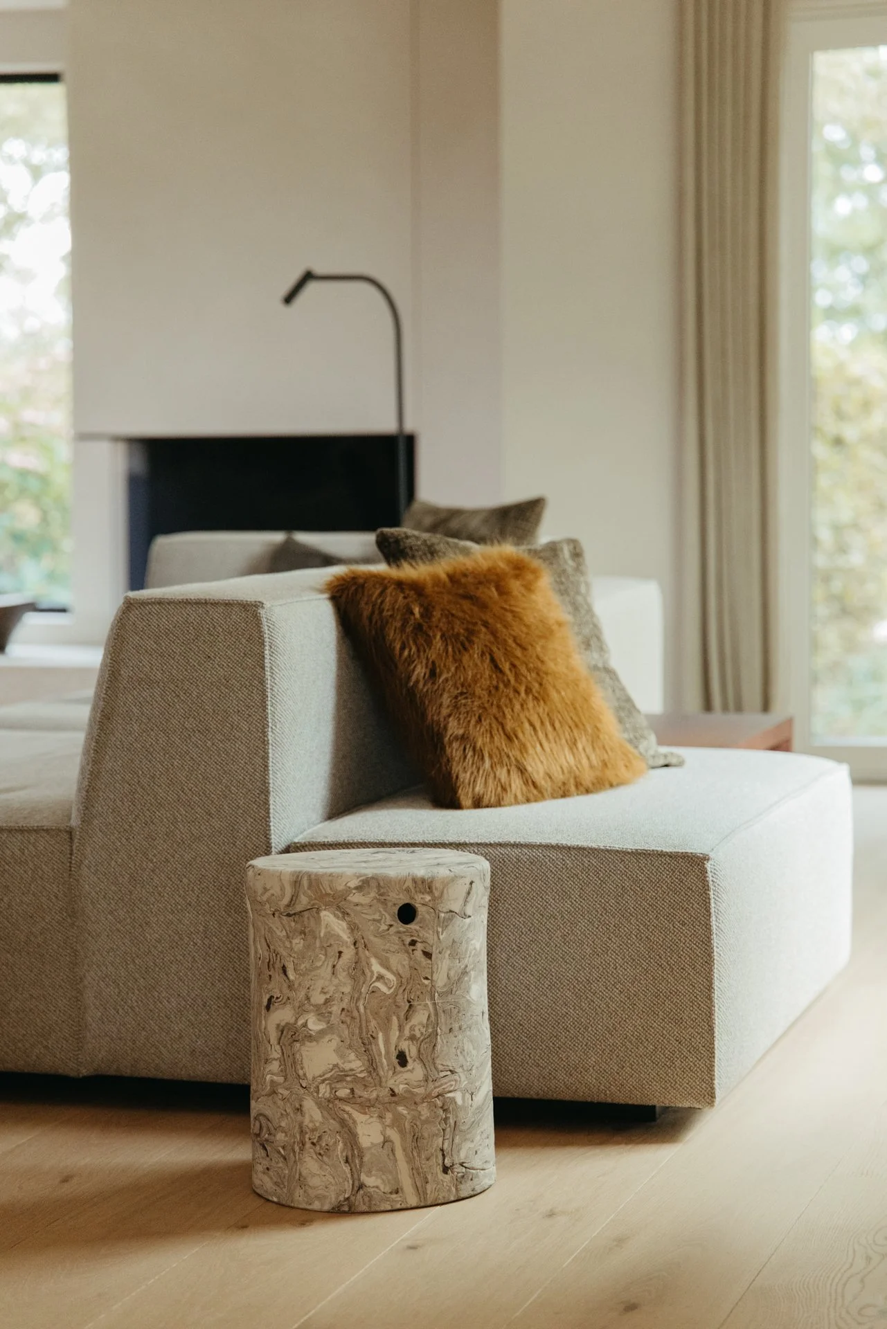 Woonkamer met beige bank en gekleurde kussens, keramische bijzettafel, houten vloer, grote ramen met gordijnen, buitenzicht met bomen.