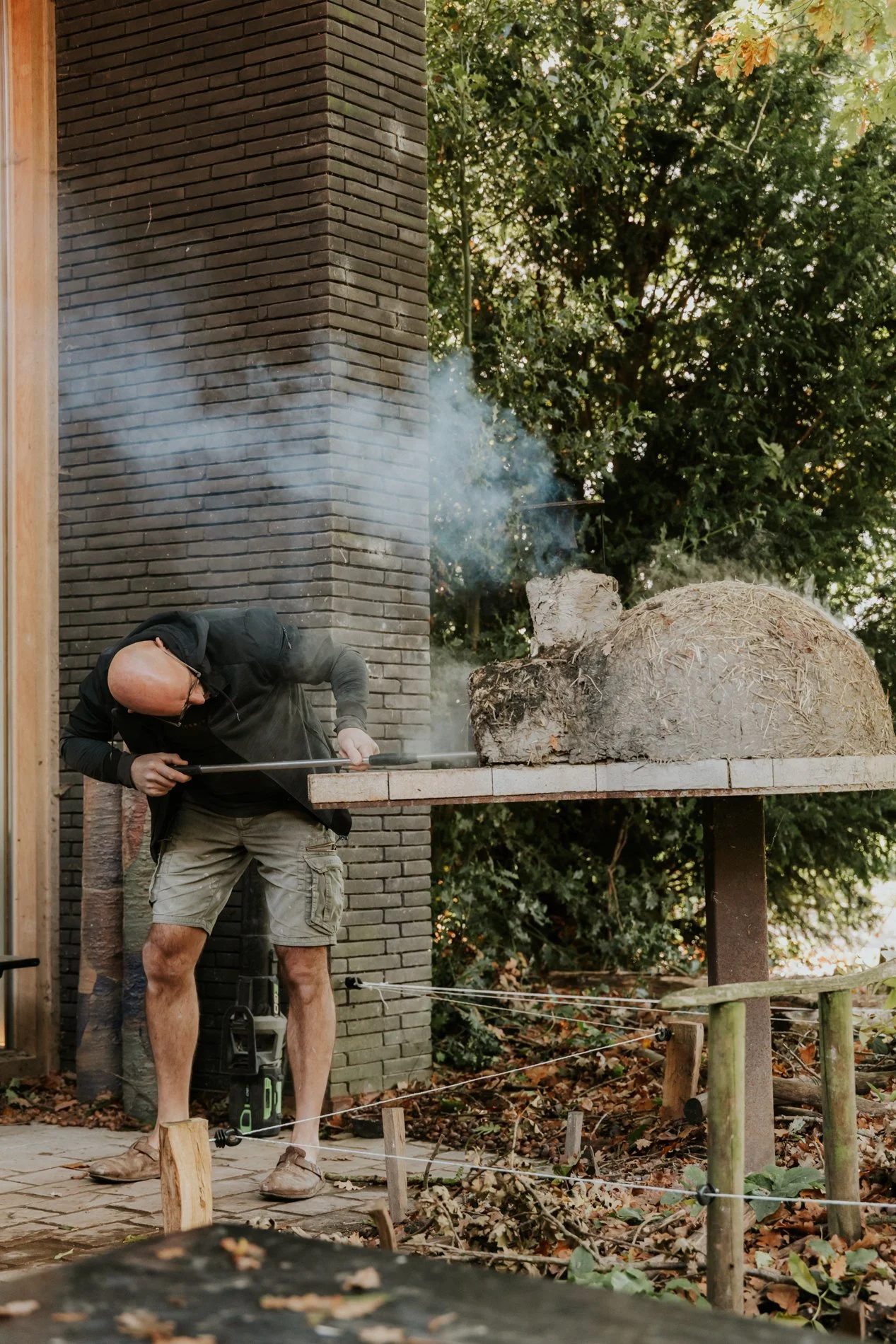 Kalende man die in de houtoven een pizza aan het afbakken is op de achtergrond de tuin en het atelier.