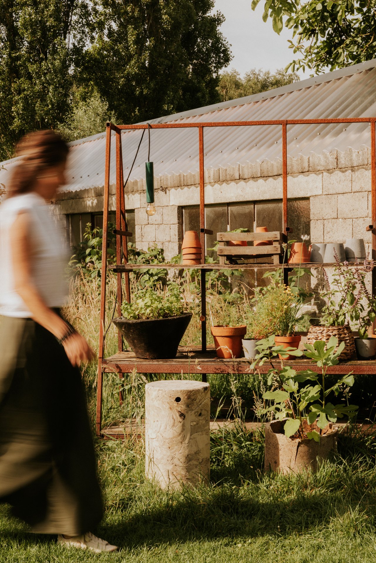 Een tuin met plantenbakken en tuinbenodigdheden, met een muur op de achtergrond en een boomgaard of bos in de verte. In de voorgrond loopt een persoon langs.