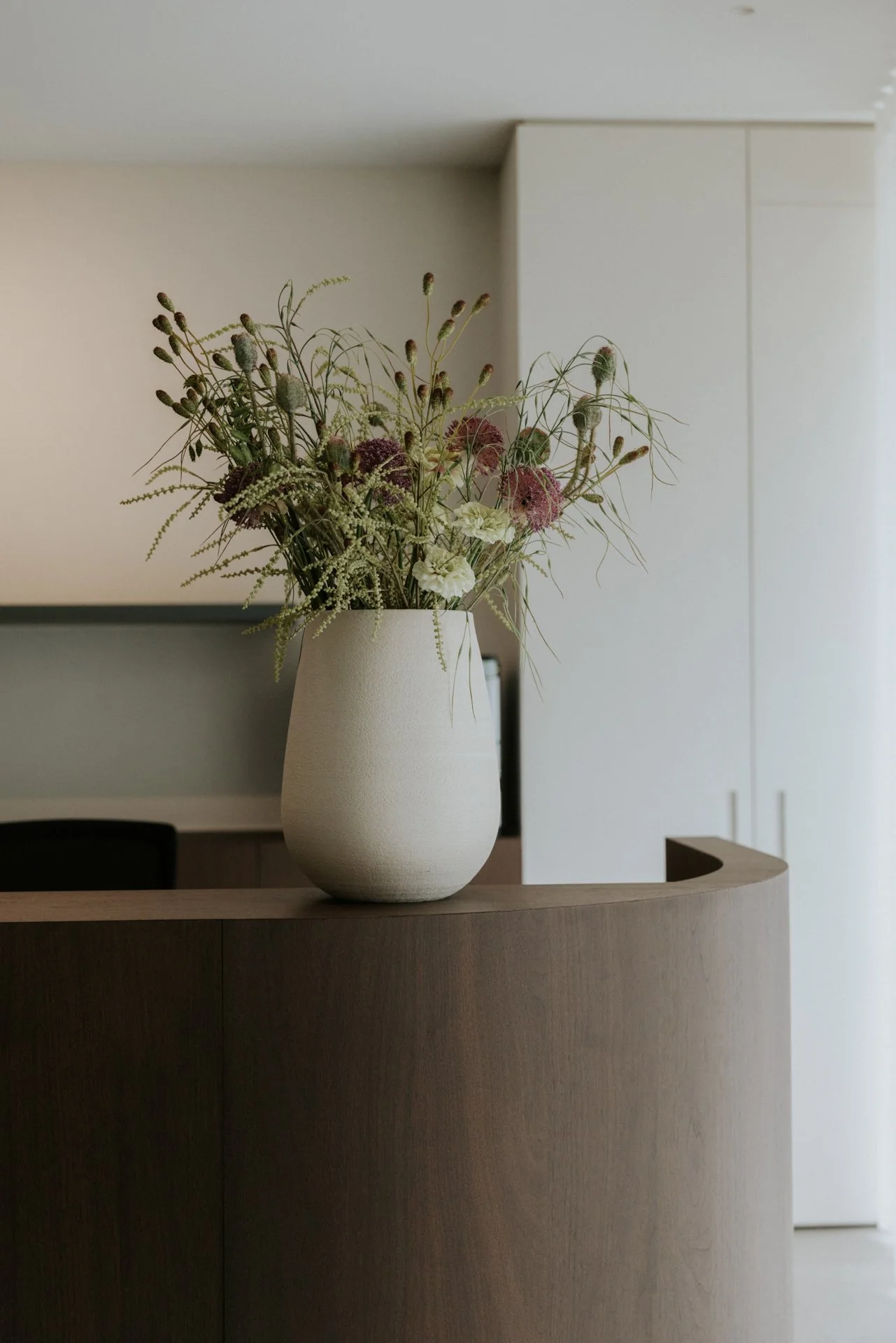 Een witte vaas met een boeket bloemen en takken op een ronde houten tafel in een interieur met witte en beige muren.