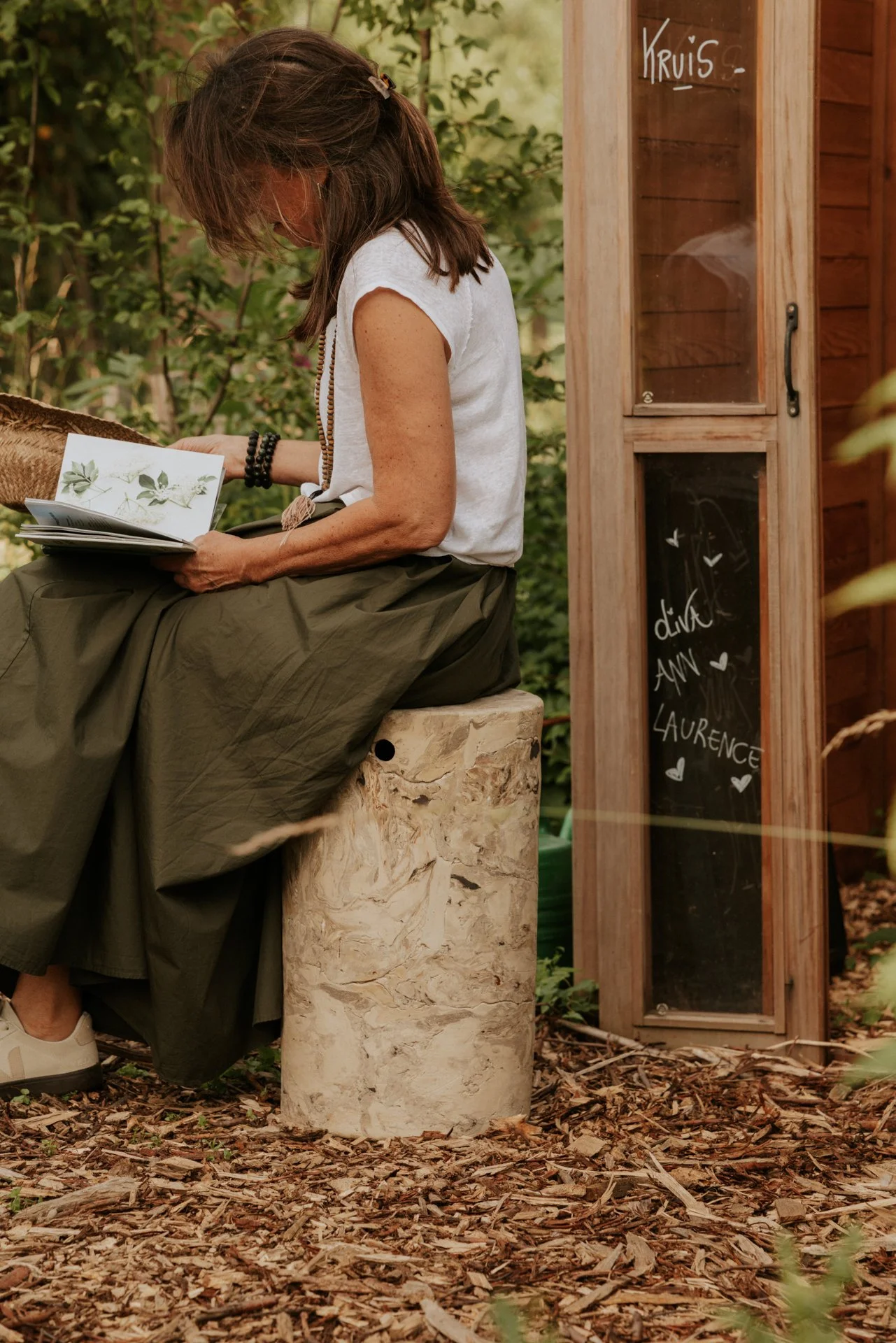 Vrouw zit op een krukjeen leest een boek in een tuin met groene planten. Een houten kast met een kleine chalkboard ernaast met schrijfletters en hartjes, waarop 'Kruis' en namen 'Anna' en 'Laureen' staan.