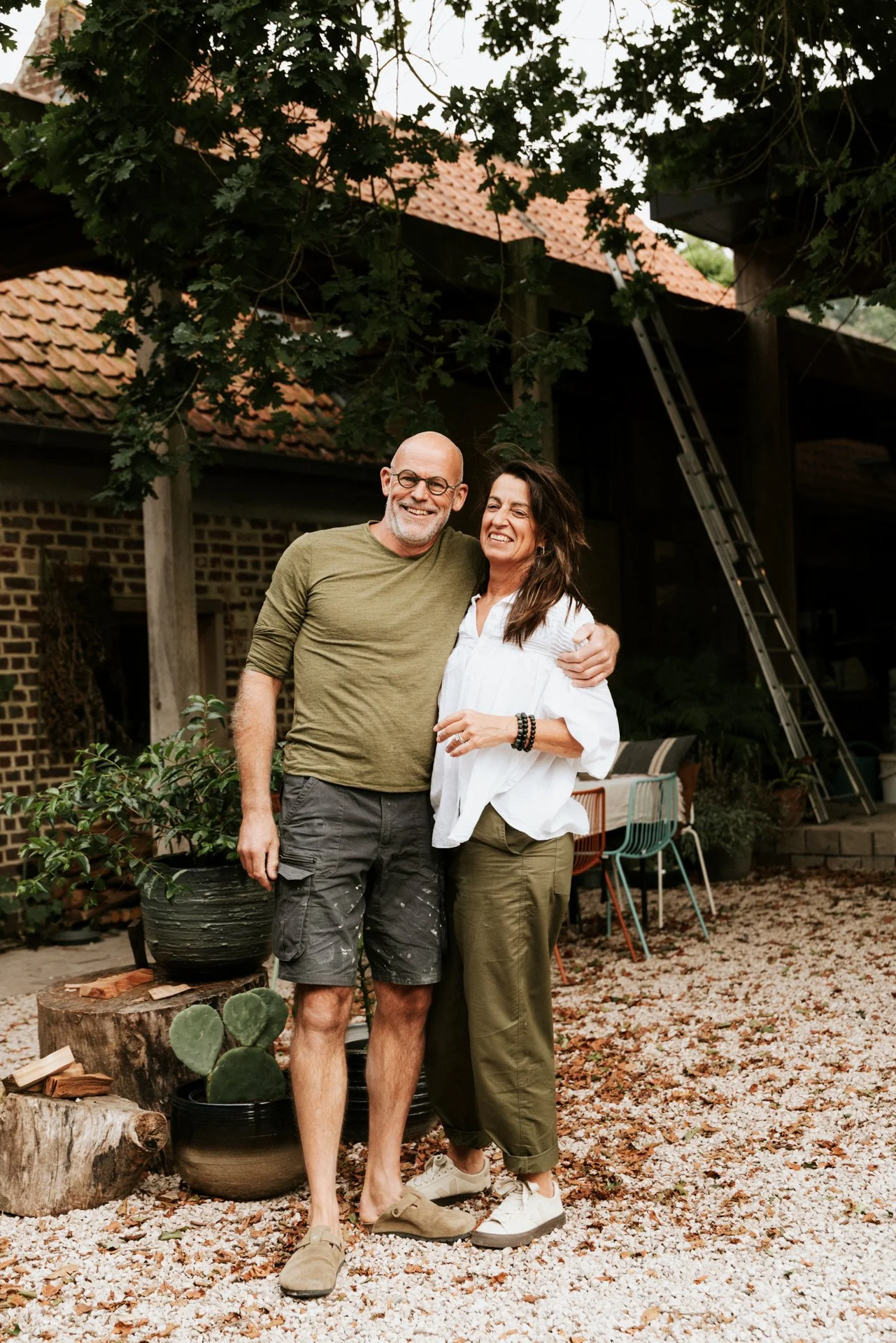Twee mensen, een man en een vrouw, staan samen buiten in een tuin, lachen en omhelzen elkaar, met een huis en tuinmeubelen op de achtergrond.