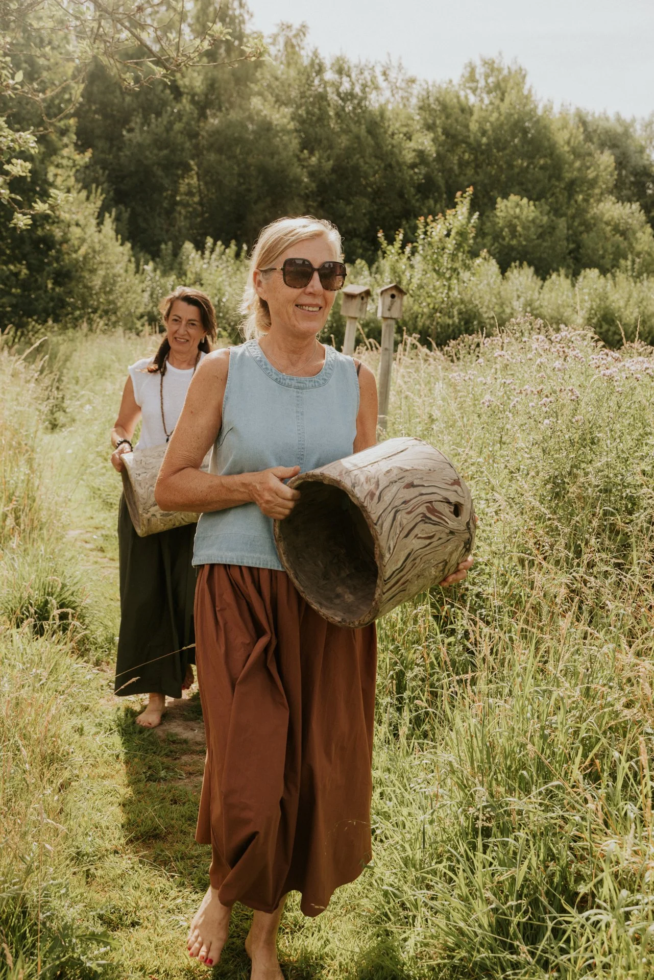 Twee vrouwen wandelen in een veld met bloemen en bomen, de voorste vrouw draagt een grote zonnebril en houdt een krukje vast, de tweede vrouw volgt op de achtergrond.