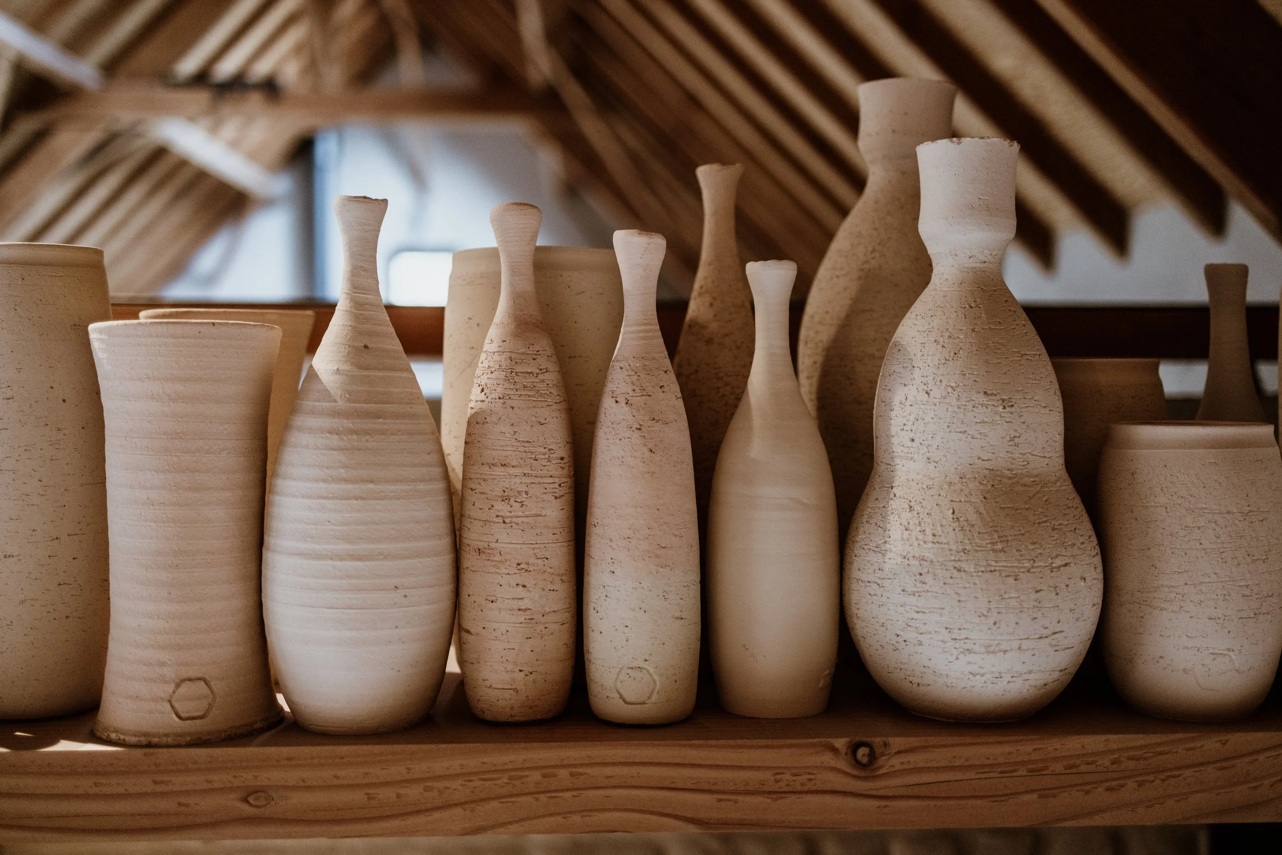 Een collectie van onbewerkt keramiekvaasjes en potten op een houten plank, binnen een ruimte met een rieten dak.