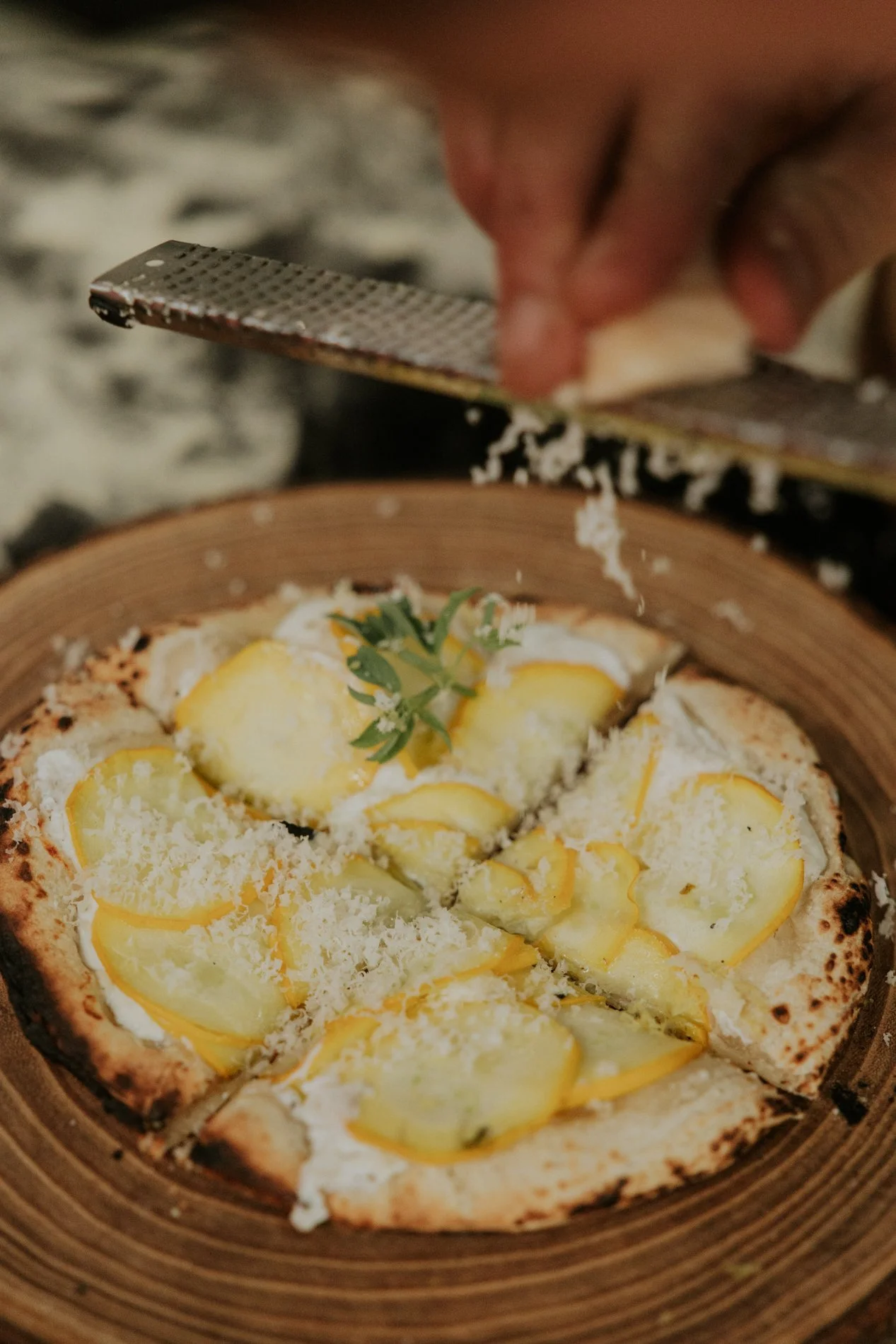 Pizza waar kaas over geraspt zal worden, hand zichtbaar, achtergrond bord in keramiek.  Atelier Pyotr Morf.