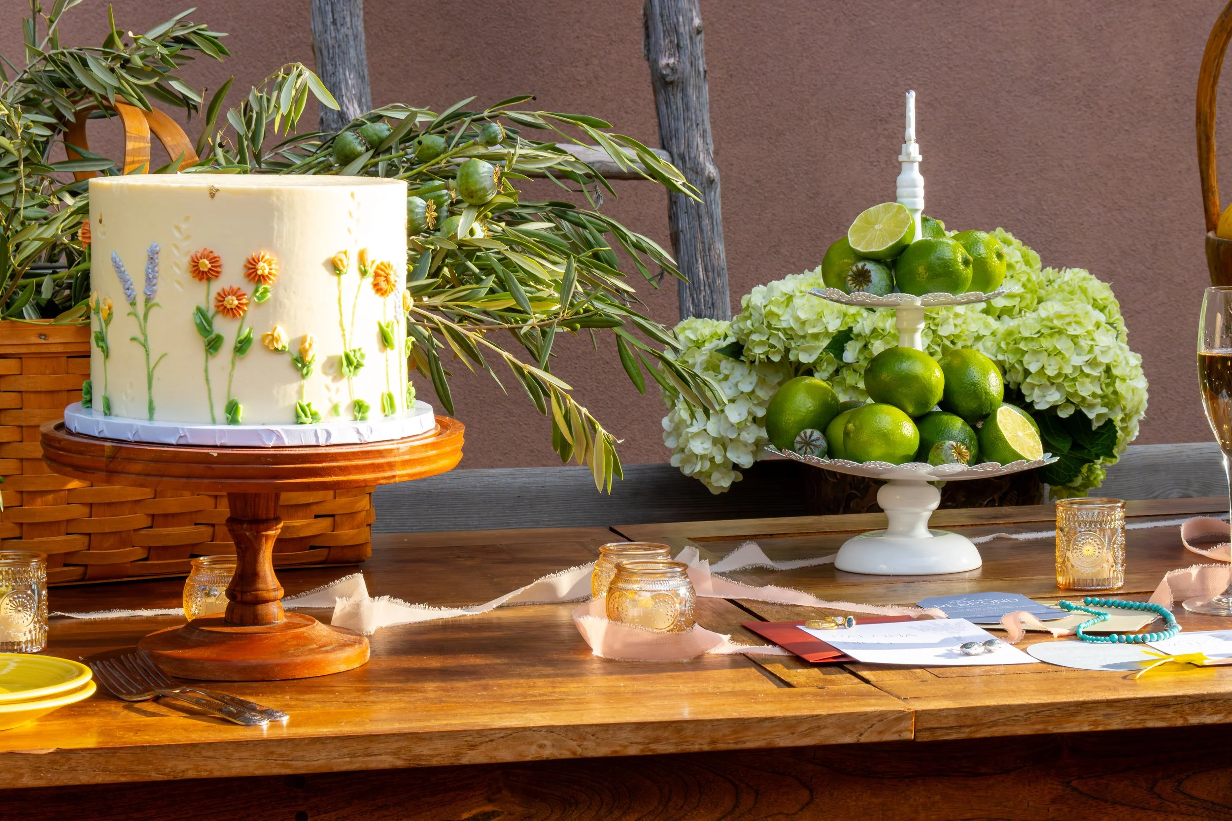 A decorated birthday cake with floral icing, a tiered tray with limes, and a floral centerpiece on a wooden table. There are paper lanterns, plates, utensils, and glasses around the table.