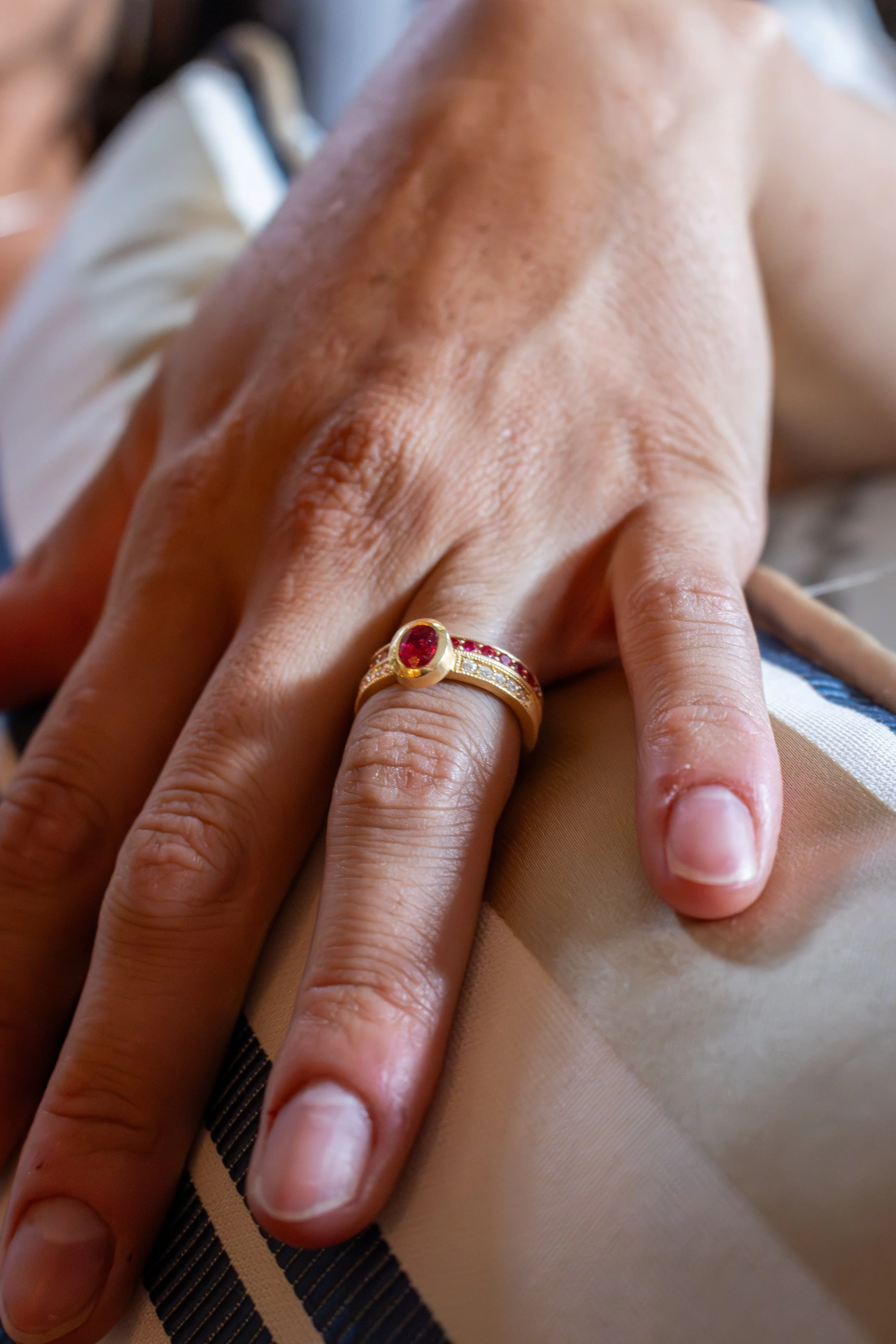 Close-up of a hand with a gold ring with red gemstone and small diamonds, resting on person's knee.