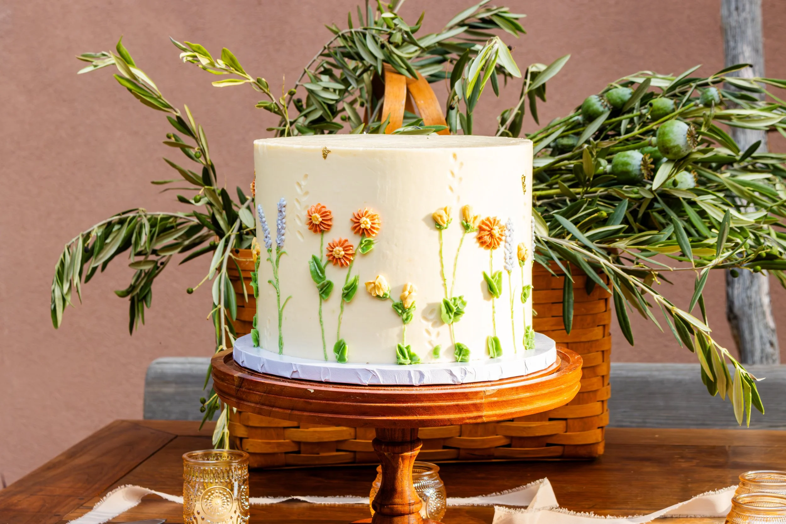 White cake decorated with colorful flowers and green leaves, placed on a wooden cake stand on a table with a small glass candle holder nearby. In the background, there is a basket with olive branches.